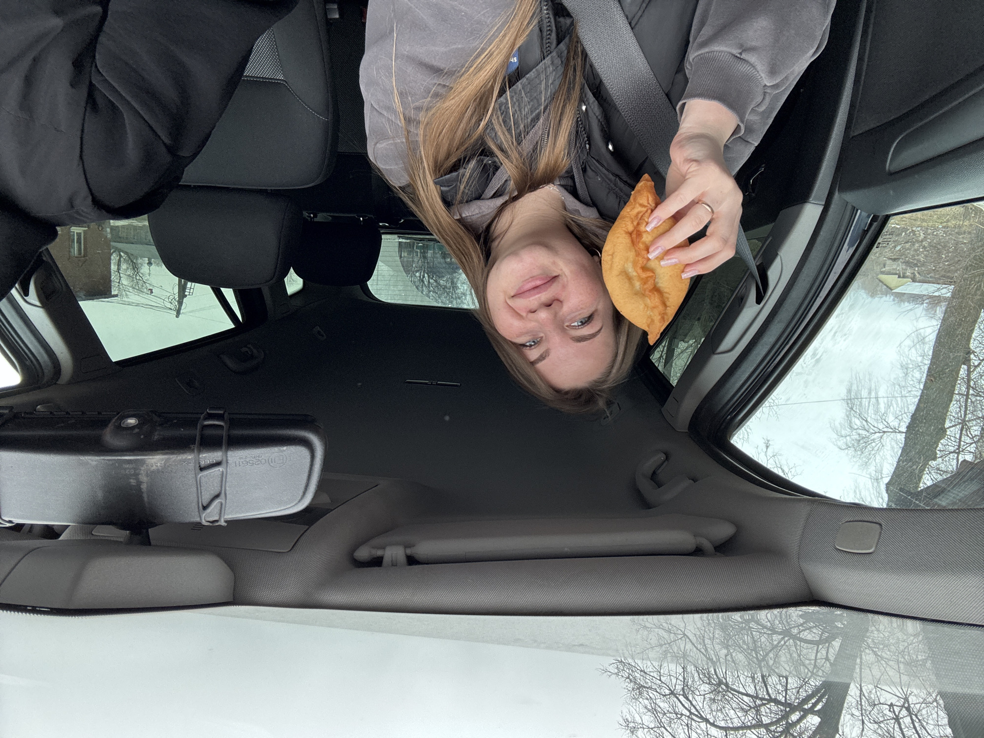 A woman sits in the driver's seat of a car, holding a fried pastry. She is wearing a grey sweatshirt over a black vest, with a seatbelt across her chest. Outside the car window, bare trees are visible, and a building with windows is seen through the back window. The rearview mirror is visible in the center of the frame. The image was taken in Фоки, Russia.