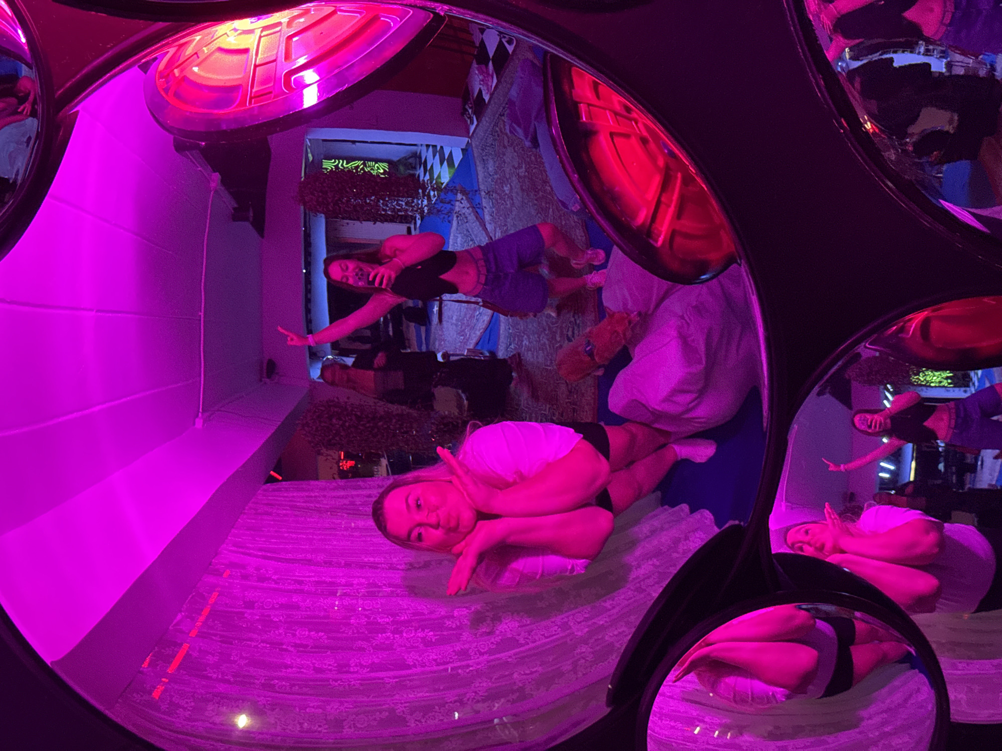 The image depicts an indoor environment illuminated by vibrant pink and red lighting, suggesting a club or entertainment venue. The scene is captured through a fisheye lens, which distorts the perspective and creates a rounded, immersive effect.
In the foreground, a woman is reflected in a large, circular mirror. She is wearing a white t-shirt and black shorts, and her hands are positioned near her face as if making a playful gesture. Her reflection is large and occupies a significant portion of the frame.
To the right, another woman is visible, also in a mirror reflection. She is wearing a black crop top and denim shorts, and she is holding a phone and posing with her arm raised. This reflection appears to be in a different mirror, positioned higher and to the right of the first.
In the background, several other people can be seen, though their features are less distinct due to the lighting and distance. The environment includes what appears to be sheer, lacy curtains or drapes illuminated by the pink light, and a patterned rug on the floor. There are also elements that suggest a stage or performance area, with some equipment and lighting fixtures visible.
The overall impression is a dynamic and playful scene, likely taken during a social gathering or event. The location of Phuket City Municipality, Thailand, is not directly evident from the visual cues in the image itself.
