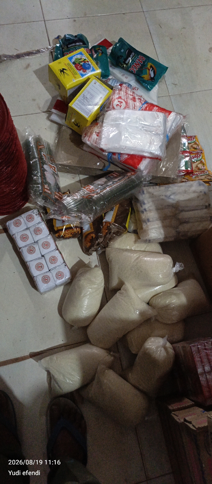 In Rawajitu Selatan, Indonesia, an overhead shot captures a cluttered floor scene with various goods. A red, circular object, possibly a basket or mat, lies on the left. Numerous plastic-wrapped packages of white granular material, likely rice, are scattered across the floor, some in larger sacks and others in smaller, more defined blocks. Several boxes, predominantly yellow and white, are present, some with visible text and graphics. Green plastic pouches with text, appearing to be food products, are also visible. Additionally, there are stacks of clear plastic cups, wrapped in plastic. A small cluster of individually wrapped white items, arranged in a grid, are on the left side of the image. The floor is tiled with light-colored squares. The bottom left corner displays a timestamp and name: "2026/08/19 11:16 Yudi efendi".