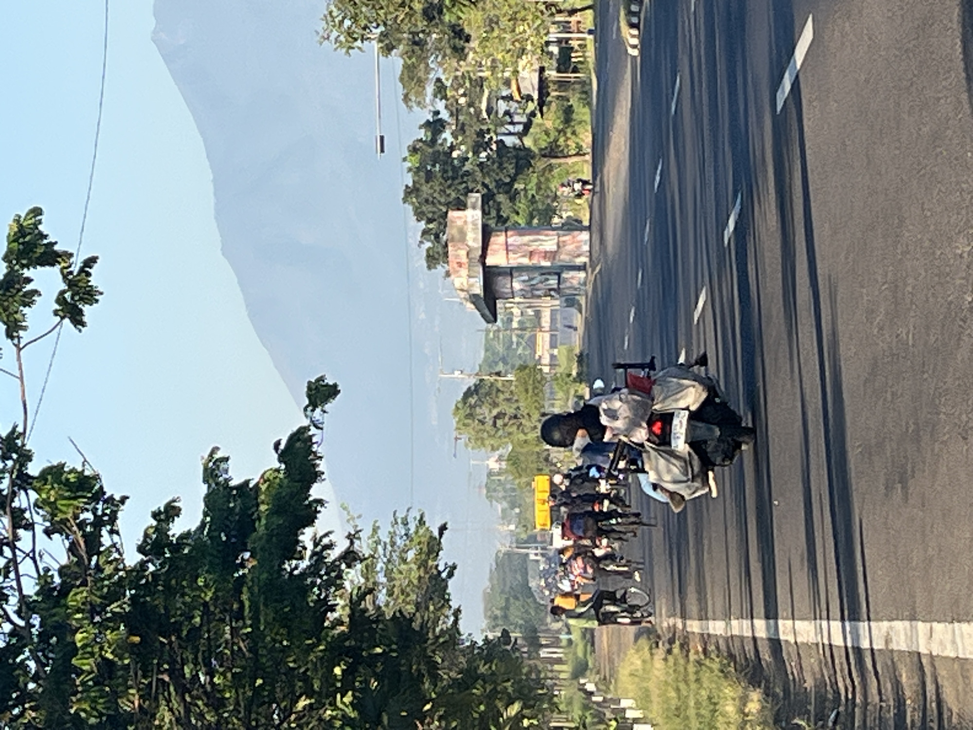 In Siring, Indonesia, a paved road is captured during daylight hours under a clear sky. A hazy mountain range dominates the background. In the foreground, a group of individuals are riding motorcycles and bicycles, some carrying luggage. A motorcycle with a rider wearing a black head covering and a light-colored jacket is in the immediate foreground, moving away from the camera. Further down the road, a cluster of cyclists are visible. To the right, an unfinished concrete structure with colorful paintwork stands beside the road, and trees line the sides of the street. The road is marked with white lines and has a curb on the left side.