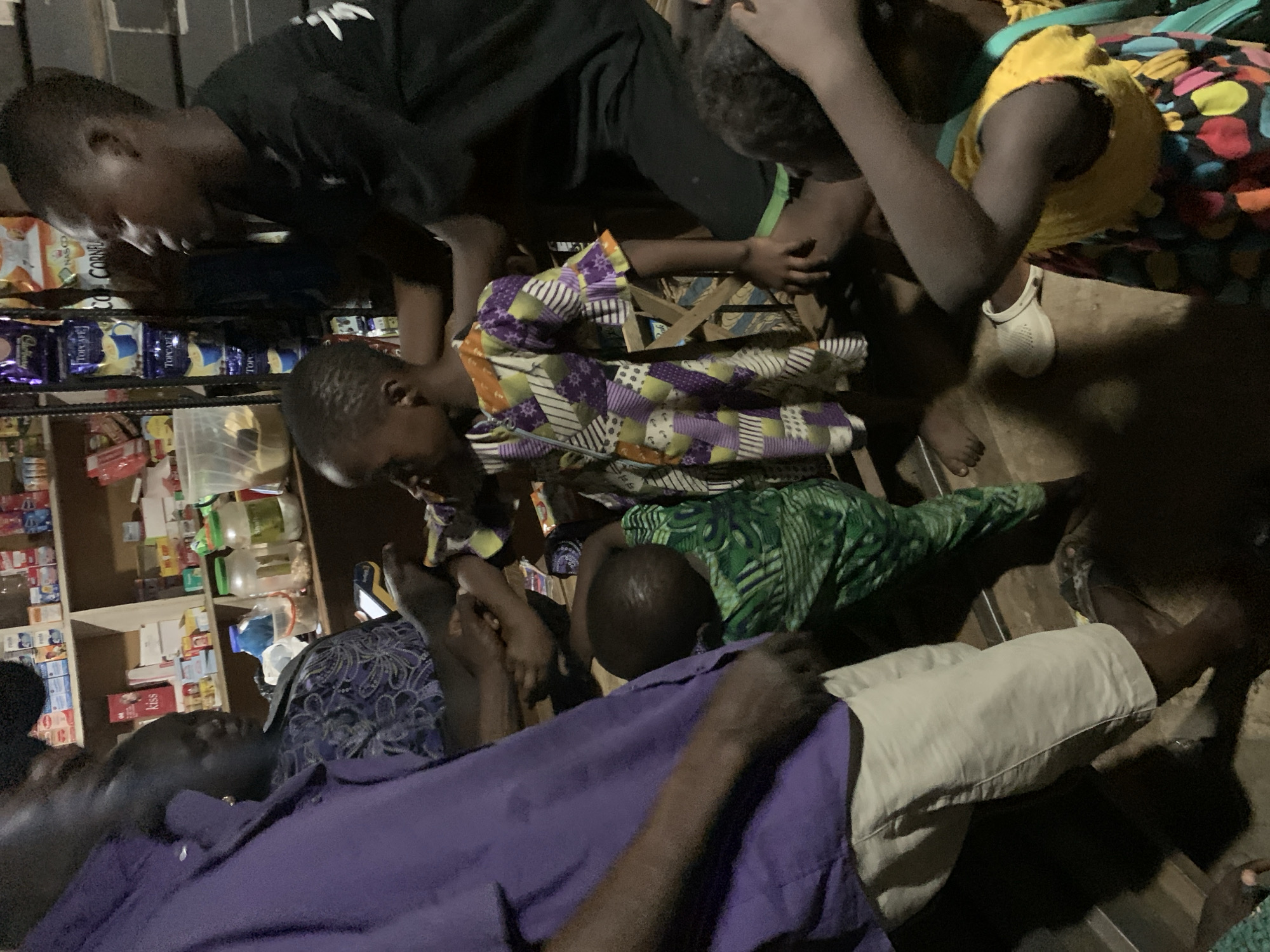 In Gbeleko, Nigeria, a group of people gather in a dimly lit, cluttered shop. Shelves stocked with packaged goods line the background. A child, wearing a yellow top with colorful polka dots and white shoes, is surrounded by others. Another child, wearing a colorful patterned top, is held in a chair. An adult, wearing a purple shirt and white shorts, is also present. The scene appears to be an intervention or gathering of concern around the child.