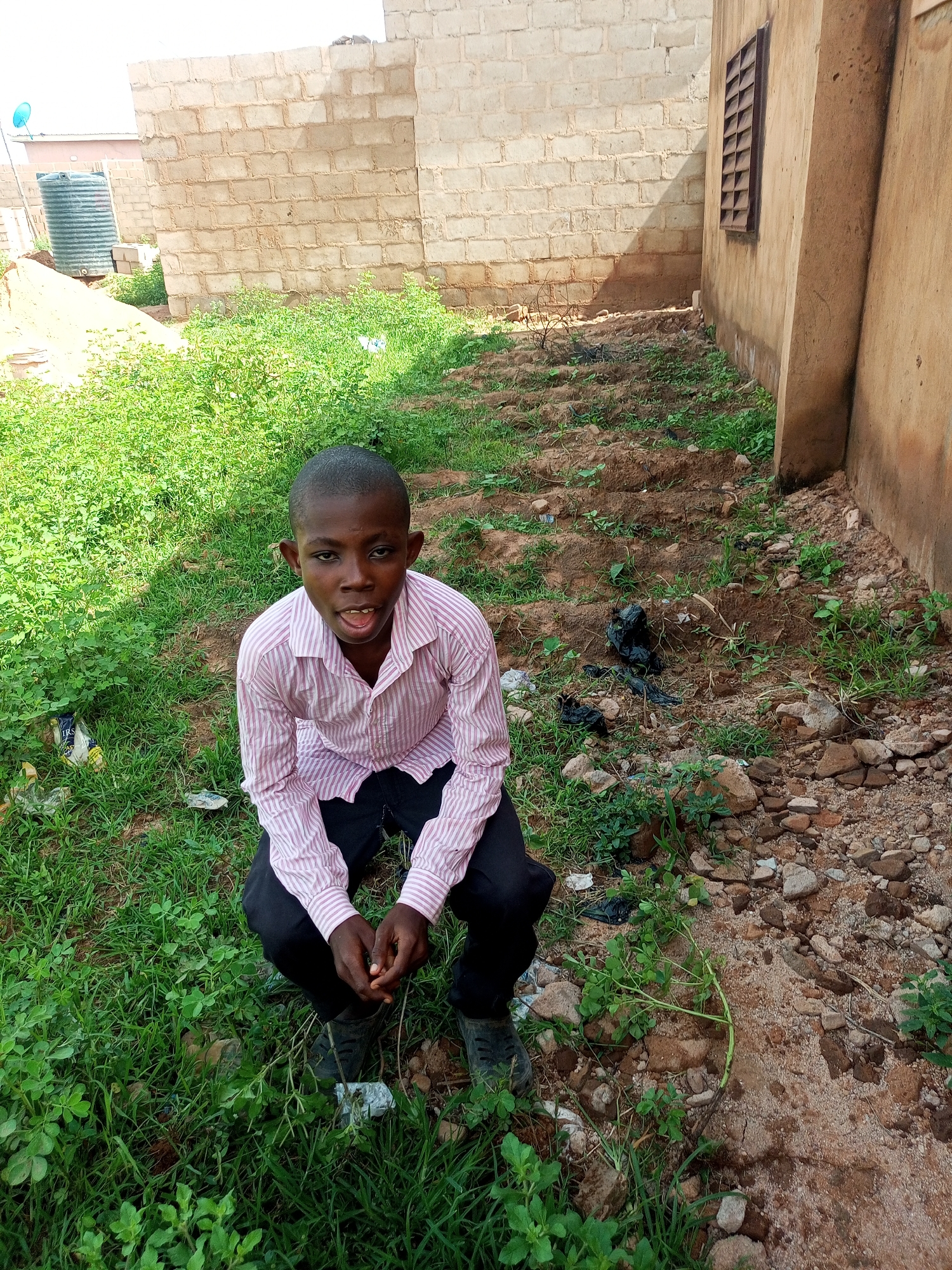 A young Black male wearing a pink and white striped button-down shirt and black pants is crouching outdoors on a patch of dirt and grass. He is looking directly at the camera with his mouth open. Behind him are two unfinished brick buildings and a large green water tank. The ground is uneven and covered with rocks and sparse vegetation. A satellite dish is visible on top of one of the buildings.