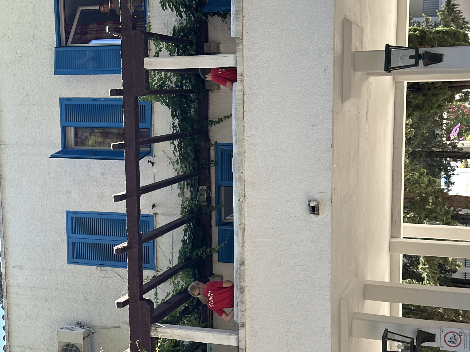This image depicts a balcony area on a building with white stucco walls and blue shutters on the windows. Two children, a girl and a boy, are visible on the balcony, both wearing red shirts. The girl, on the left, has blonde hair in a ponytail and is facing away from the camera. The boy, on the right, is also facing away from the camera. In the background, a person is looking out of a window. Below the balcony, there is a covered walkway with white pillars and lampposts. Trees and foliage are visible behind the walkway. The setting appears to be warm and sunny. The building's architecture, including the stucco walls and blue shutters, is characteristic of Mediterranean or coastal regions. Specifically, this scene is located in Budva, Montenegro, a popular tourist destination known for its picturesque coastline and historic architecture.