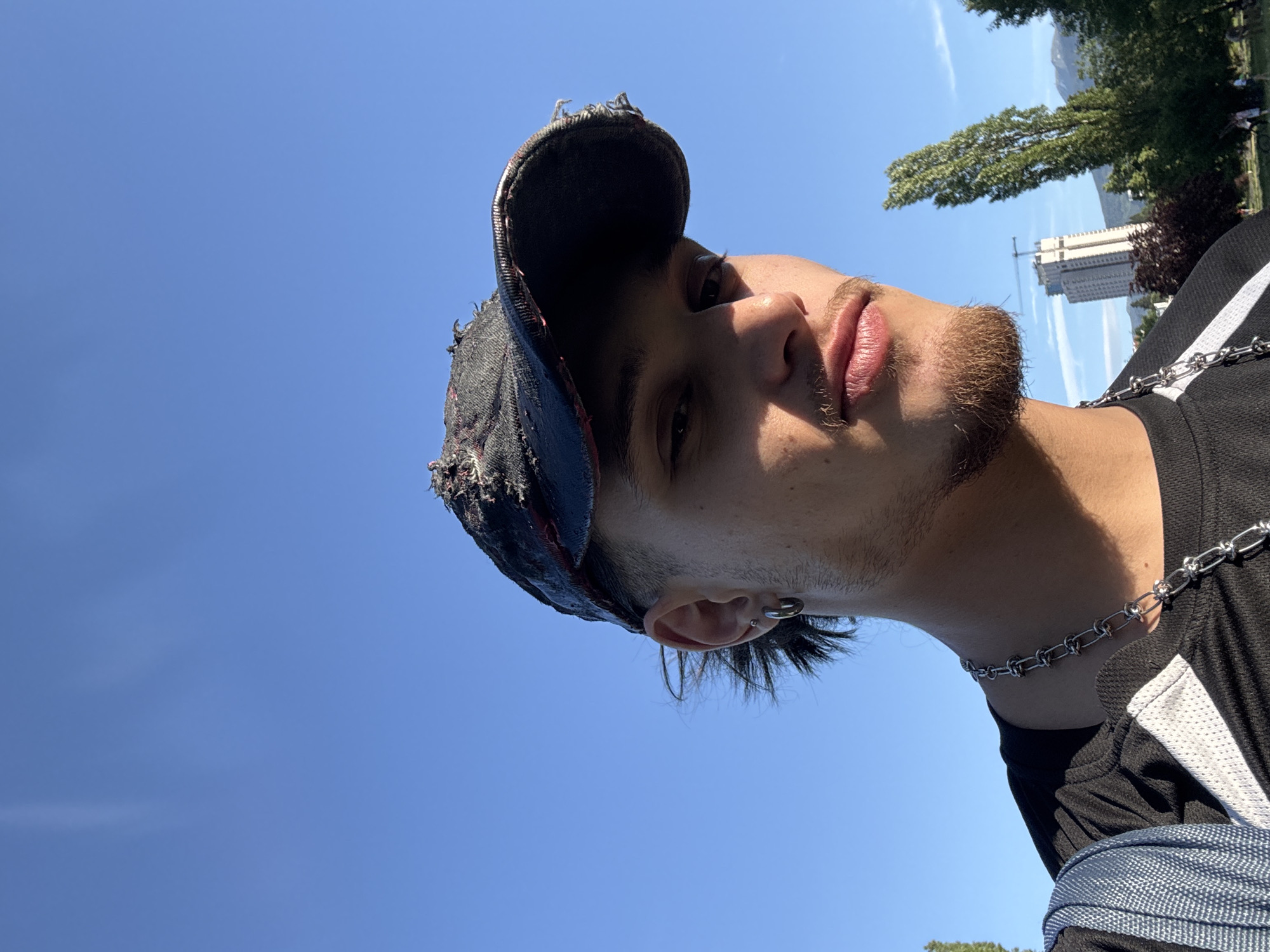 A close-up selfie of a young man with light brown skin, a patchy beard, and dark hair, wearing a distressed black baseball cap, taken outdoors on a sunny day. He is wearing a silver chain necklace and a black shirt with white accents. The background consists of a clear blue sky and trees, and a tall white building with a construction crane, suggesting an urban environment, possibly in Sofia, Bulgaria.