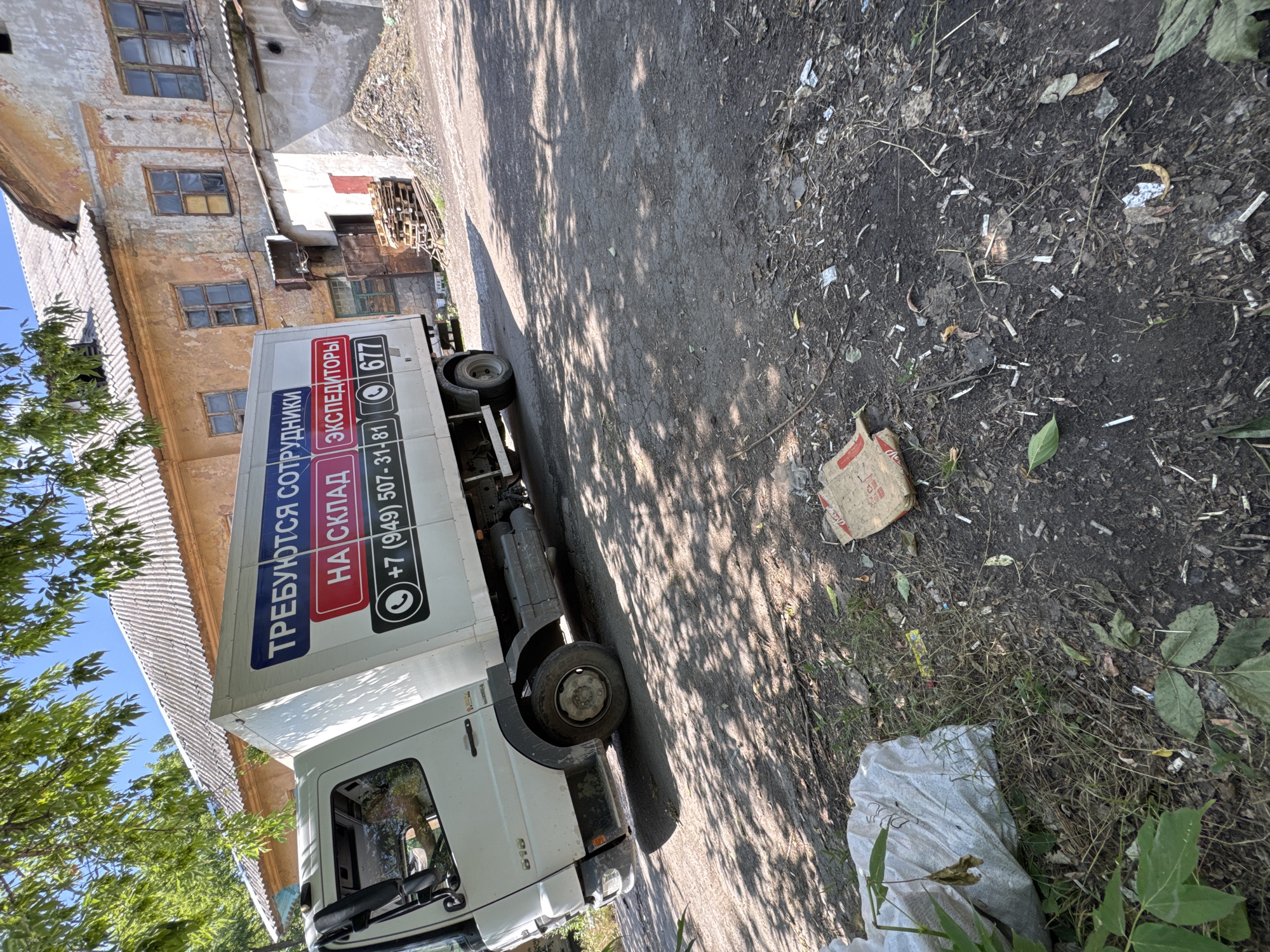 A white truck is parked on a dirt road next to an old building. The truck has writing on its side that says "ТРЕБУЮТСЯ СОТРУДНИКИ НА СКЛАД ЭКСПЕДИТОРЫ" which translates to "STAFF NEEDED FOR WAREHOUSE EXPEDITORS". Below this, there is a phone number "+7 (949) 507-31-81" and a partial number "677" with icons of a phone and a mailbox. The building appears to be in disrepair, with peeling paint and broken windows. The ground is covered in dirt, debris, and some patches of grass. There is a large white plastic bag on the left side of the image and a cardboard box in the center. The scene appears to be outdoors in a somewhat neglected industrial or rural area.