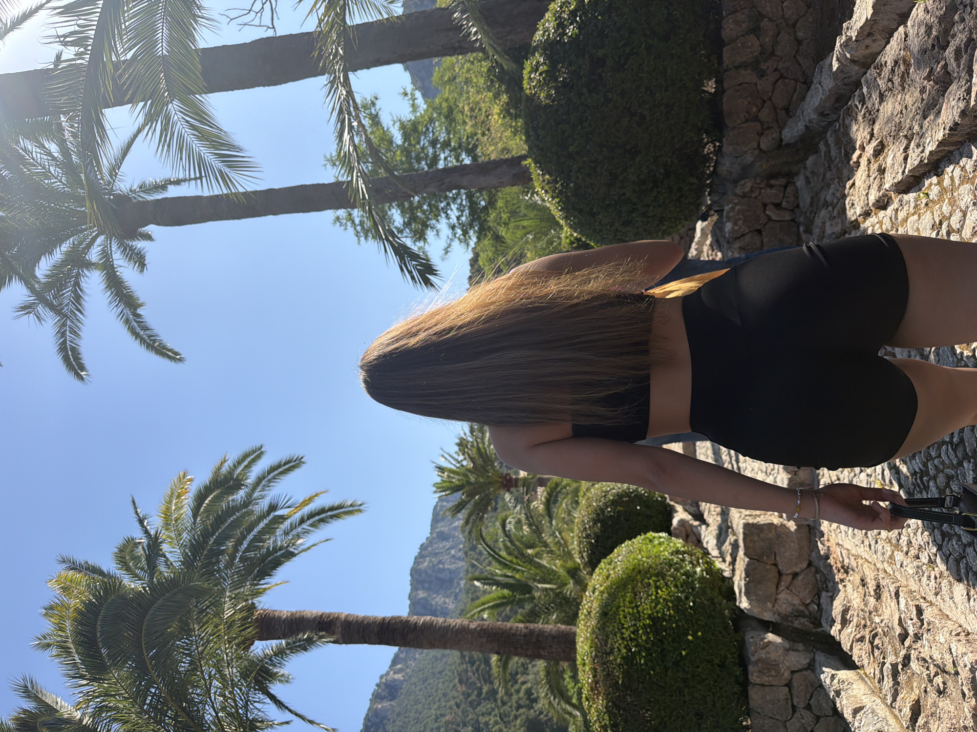 A waist-up view of a woman with long brown hair is captured outdoors on a sunny day. She is wearing a black crop top and black shorts, standing on a stone path with a purse in her hand. The environment includes lush greenery with round bushes, palm trees, and a mountain visible in the background. The setting suggests a location in Bunyola, Spain, where such natural landscapes are common.