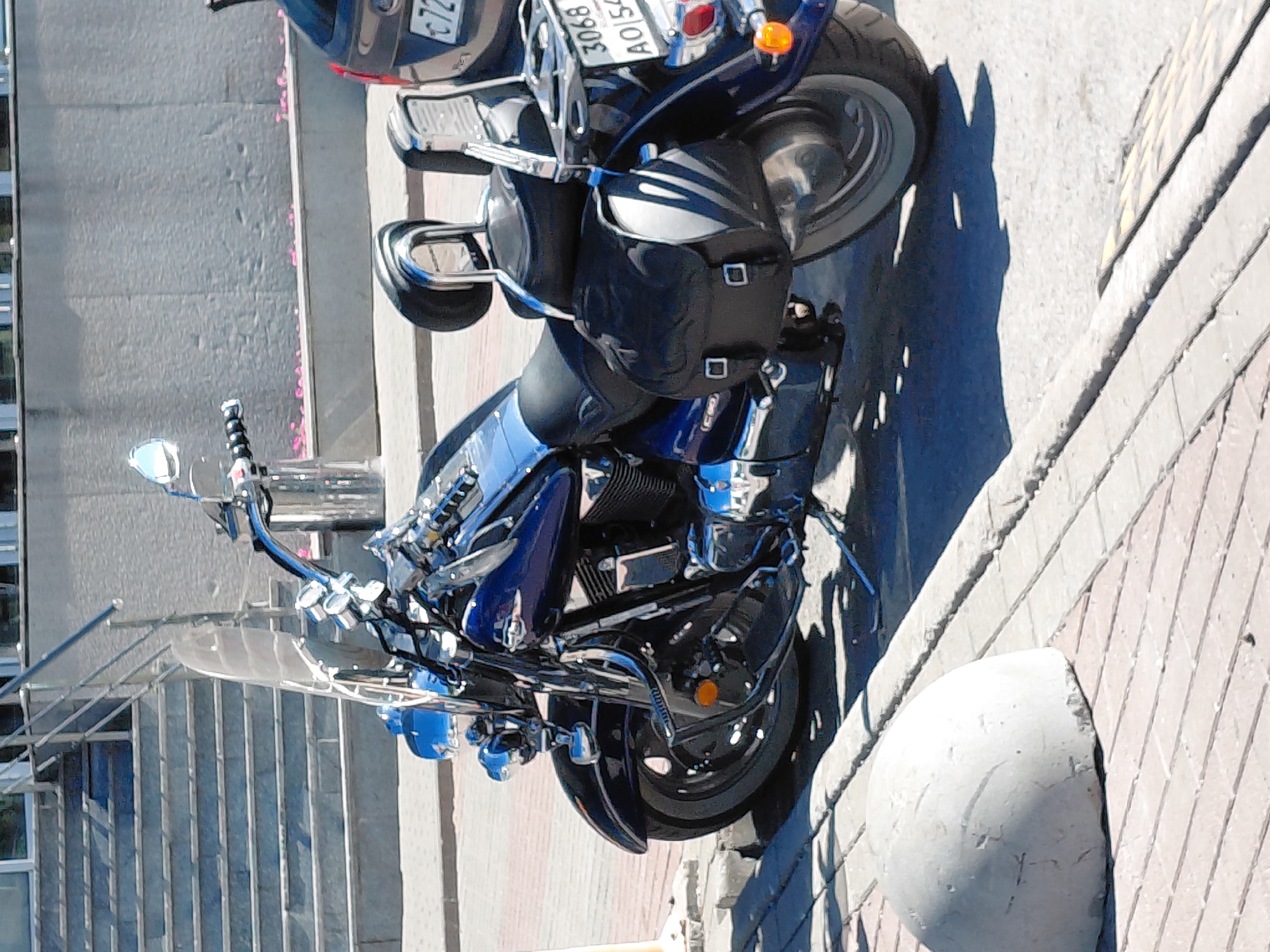 The image captures a parked blue and chrome motorcycle on a paved surface, likely a sidewalk or street, in front of a building with a stone facade and a set of stairs. The motorcycle is a cruiser-style bike with saddlebags and a windshield. A dark-colored car with a license plate that reads "3068 AOI 54" is visible to the right of the motorcycle. A concrete bollard is situated on the left side of the image, in front of the motorcycle. The scene appears to be outdoors on a sunny day, with shadows cast by the motorcycle and other objects.
