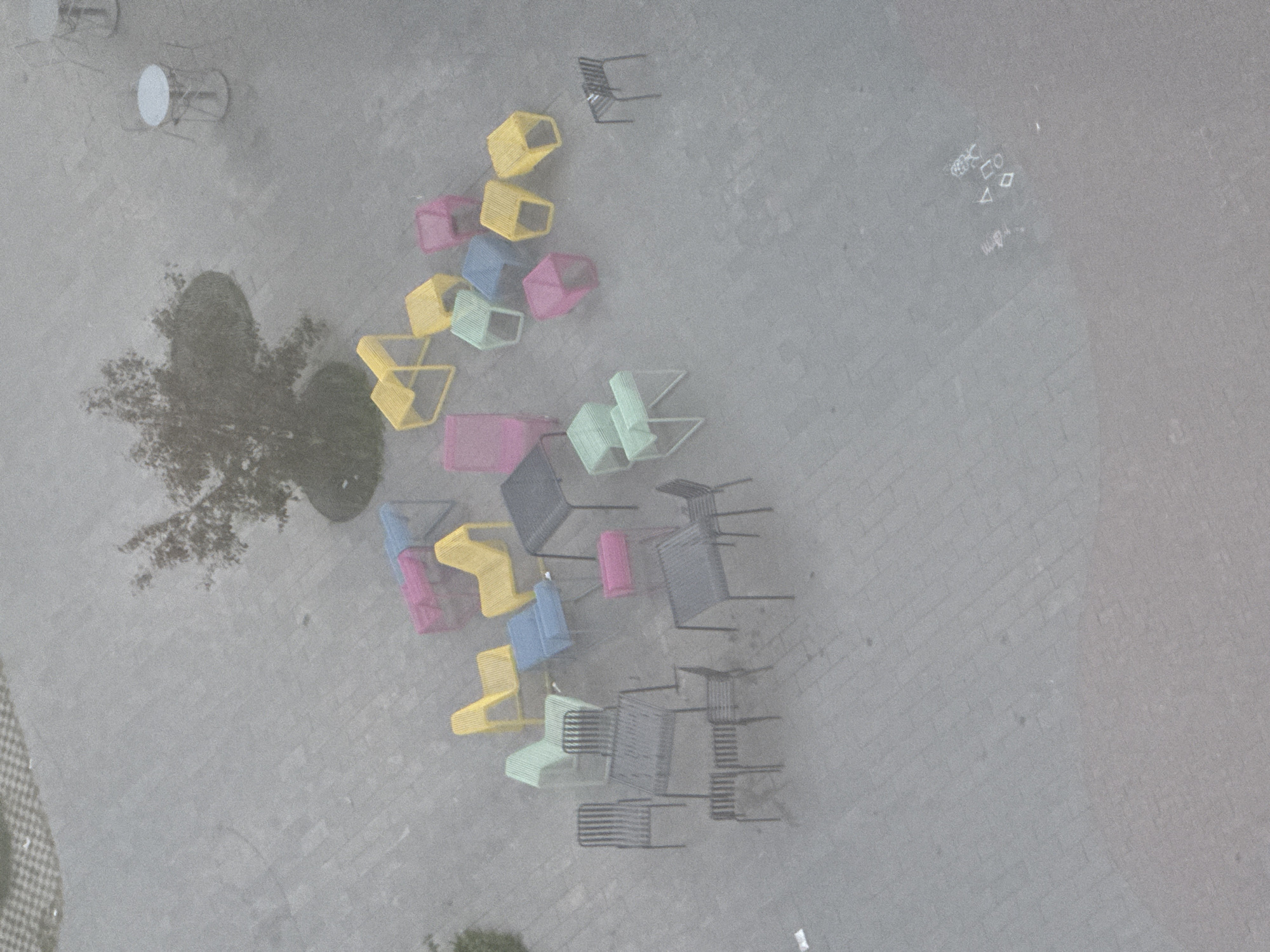 The image shows an aerial view of an outdoor space in Izhevsk, Russia. The ground is paved with gray bricks. A tree and a round table with chairs are located in the upper left corner. There are numerous chairs scattered throughout the scene, with a concentration of colorful chairs in the center-left, and gray chairs in the lower-right. Some graffiti is visible in the upper-right quadrant.