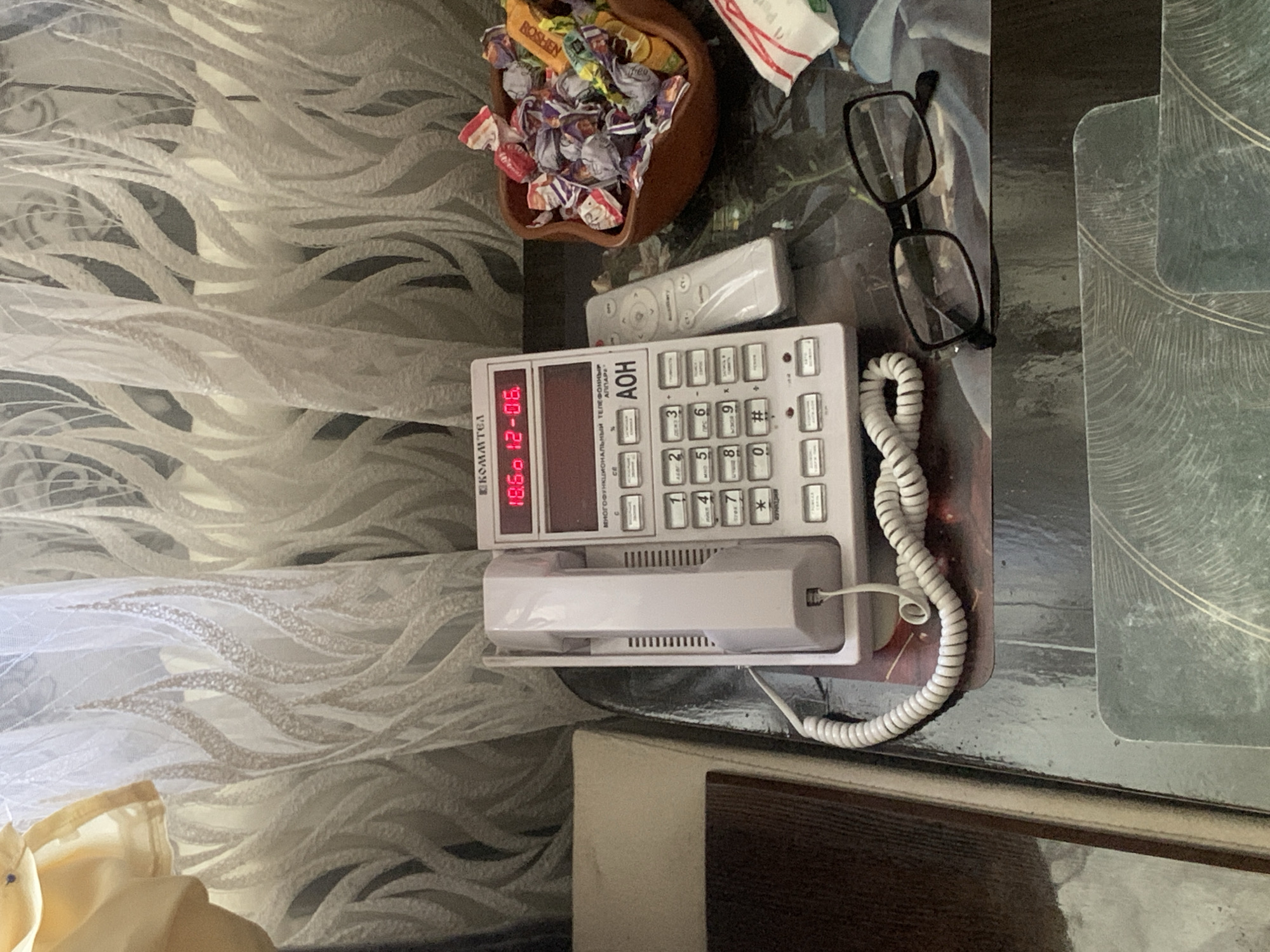 The image shows a close-up of a table with a landline telephone, a remote control, a bowl of candies, and a pair of eyeglasses. The telephone is pink and has a digital display showing "18.54 12-06". The remote control is white and appears to be for the telephone. The bowl of candies is made of dark wood and is filled with assorted wrapped candies. The eyeglasses are black-rimmed. In the background, there is a patterned curtain. The scene appears to be indoors. The lighting suggests it is daytime. The image is taken from a slightly elevated angle, looking down at the table. There are no people or animals visible in the image. The location is not specified in the image itself.