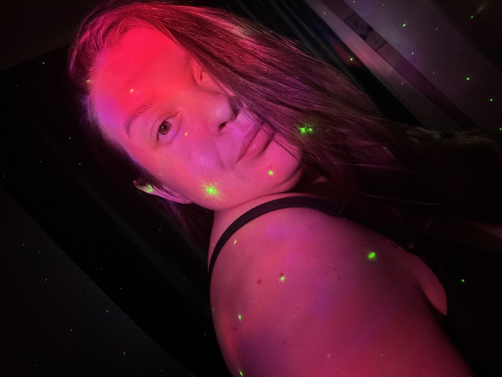 A woman is illuminated by pink and red light with green light dots on her face and shoulder. The background is dark. The location cannot be determined to be Марковский, Russia from the image.