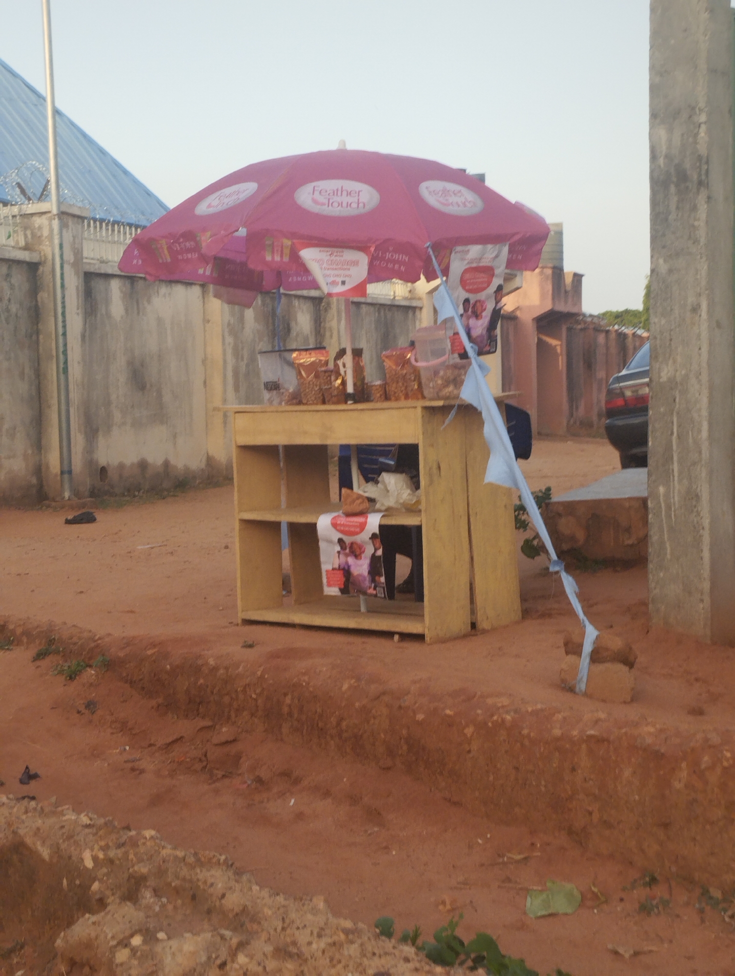 The image depicts a small roadside vendor in Tumfure, Nigeria. The vendor has set up a makeshift wooden stand under a pink "Feather Touch" branded umbrella. Various bagged goods, likely snacks or food items, are displayed on the stand. A blue cloth is tied to the umbrella support and secured to bricks on the ground. The ground is reddish-brown soil. In the background, there are buildings, a barbed wire fence, and a parked car. A concrete pillar is visible on the right side of the frame.