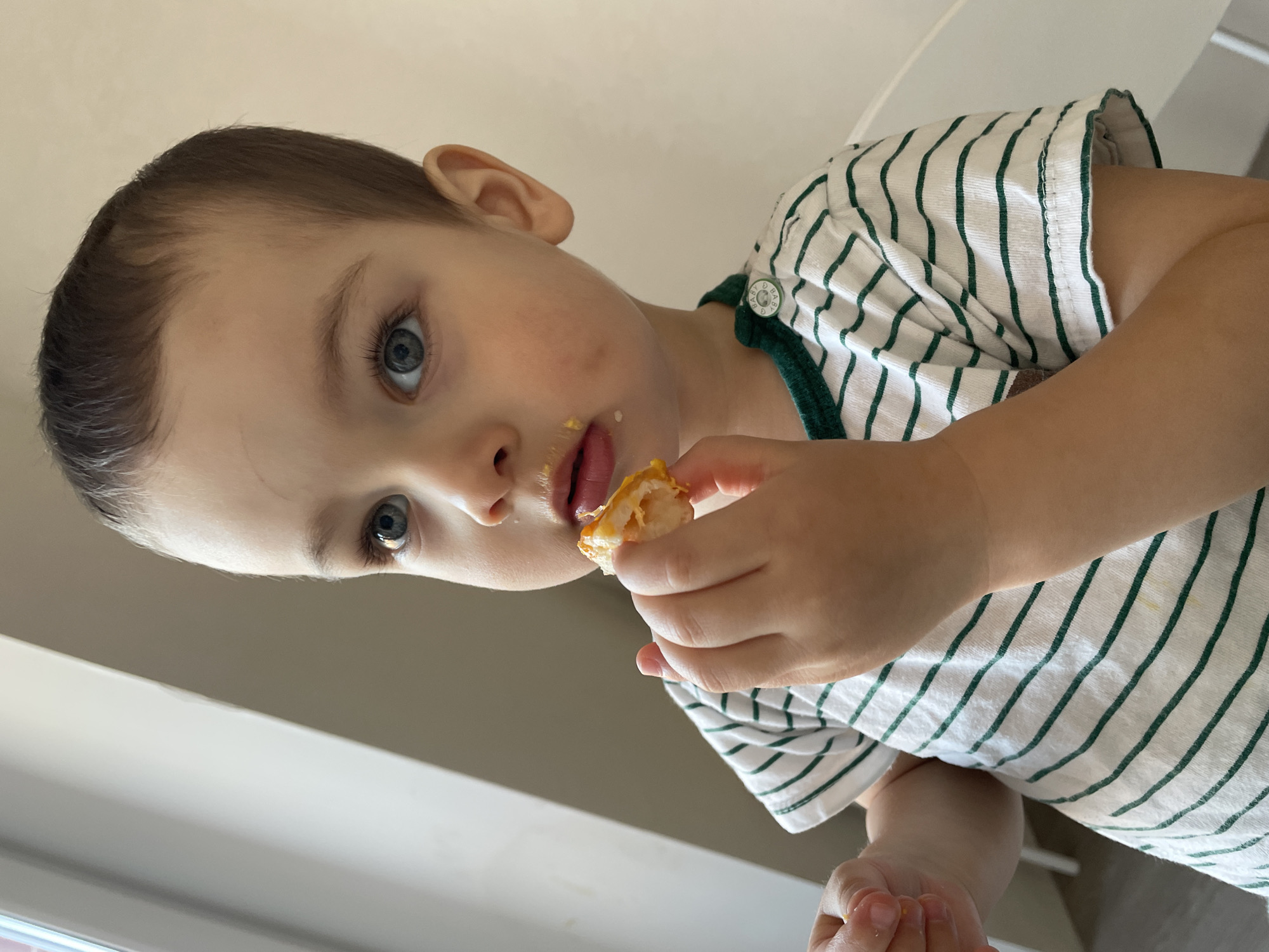 The image captures a young child, appearing to be male, in an indoor setting. The subject possesses short brown hair, blue eyes, and a minor scratch mark on the forehead. Attire consists of a white short-sleeved t-shirt featuring thin dark green horizontal stripes, a green collar trim, and two white buttons on the left shoulder, one bearing the word 'BABY'. The child holds a piece of yellowish food in the right hand and a smaller fragment in the left. Food residue is present around the mouth, consistent with an active feeding state. The subject's gaze is directed upwards and to the left of the frame. The background comprises a vertical window frame on the left, which is a source of natural illumination, and a plain, light-colored wall on the right. A white, undefined object is partially visible in the lower right background. There is no visual information within the image to ascertain its location in Izhevsk, Russia.