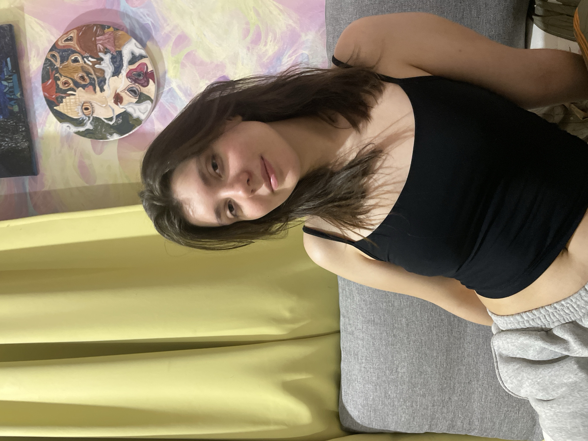 A young woman with shoulder-length dark hair and fair skin sits on a gray couch. She is wearing a black tank top and light gray sweatpants. She is looking towards the camera with a slight smile. Behind her, yellow curtains hang to the left, and a colorful abstract painting and a smaller framed artwork are displayed on the wall to the right. The room appears to be a living space within an apartment in the residential complex "Borisoglebskoye" in Russia.