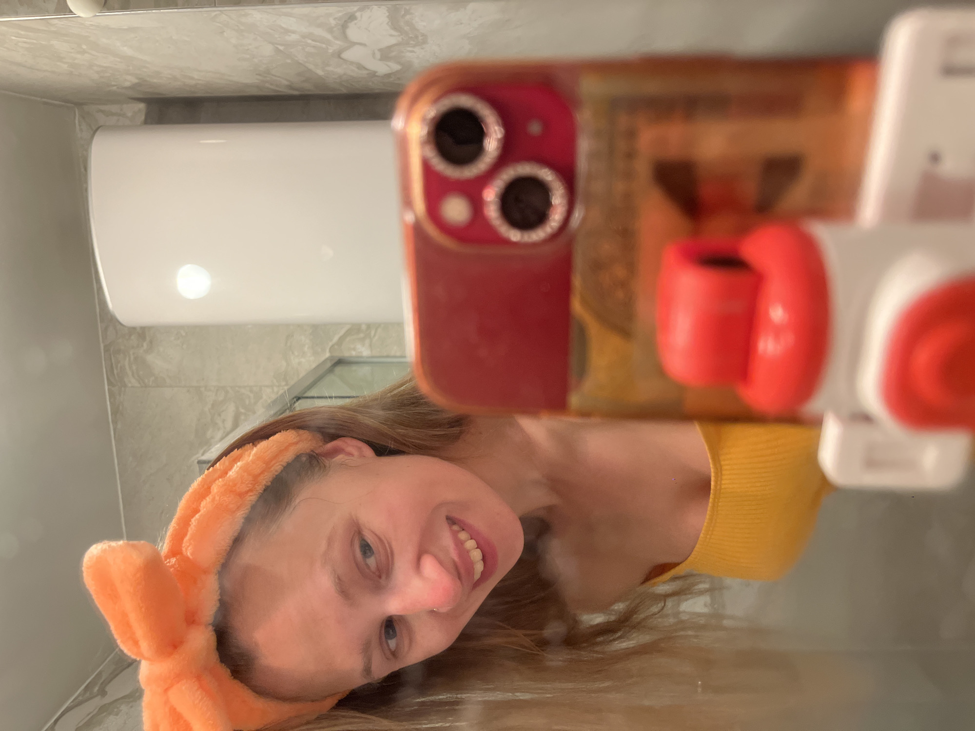 A woman is taking a selfie in a bathroom, likely in a residential setting. She is wearing an orange headband and a yellow top. The reflection of her phone is visible in the foreground. In the background, there is a white water heater mounted on the wall. The location is not discernible from the image, although the request mentions Bečići, Montenegro. However, there is no visual information to confirm this location.