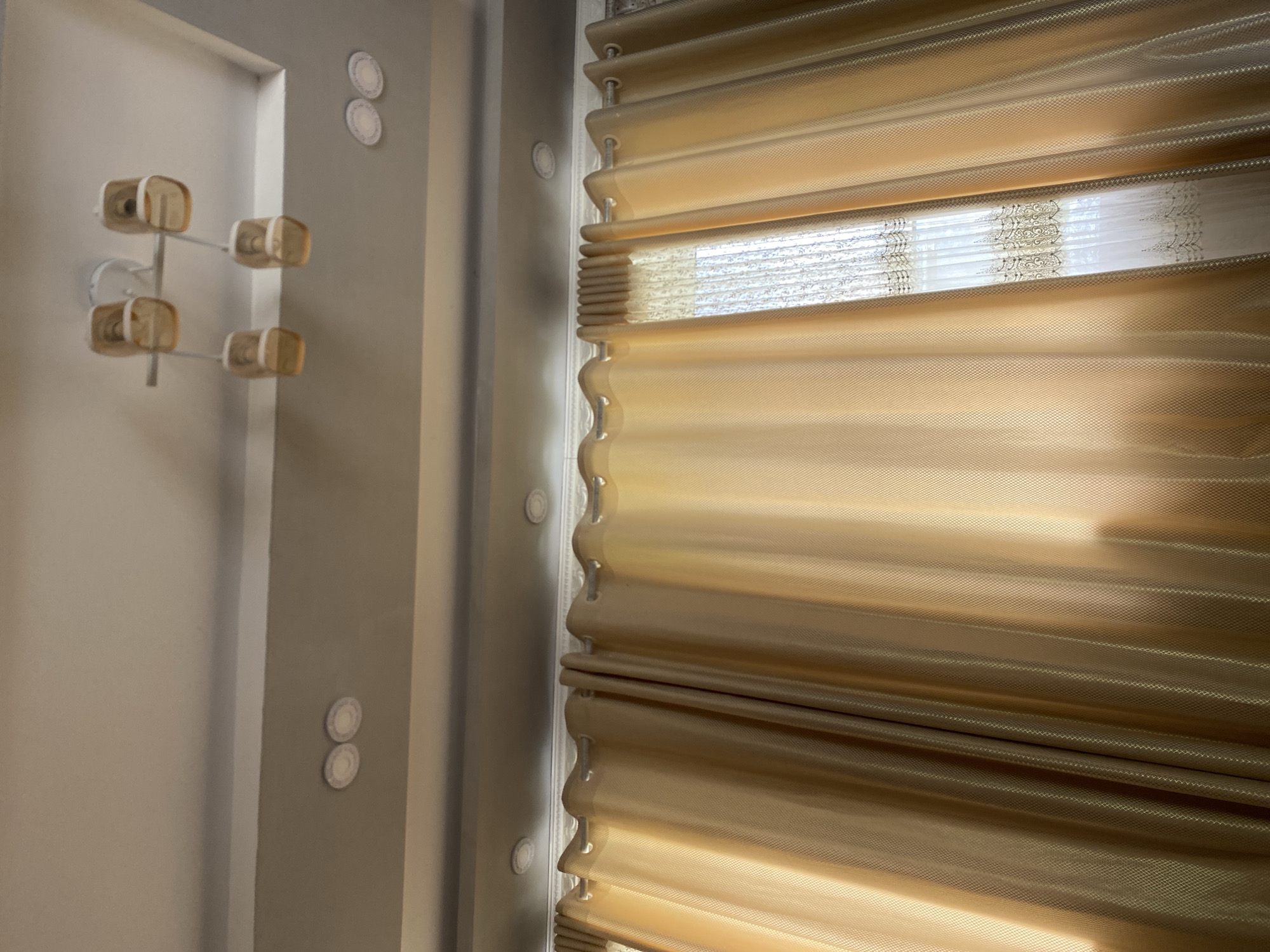The image captures an interior scene, looking up towards a ceiling and a window draped with curtains. The ceiling is painted white and features recessed lighting fixtures. A modern light fixture with four amber glass shades hangs from the ceiling. The window is covered by two layers of curtains: sheer white lace curtains with decorative trim are layered behind heavier, golden-brown curtains. Sunlight filters through the sheer curtains, casting a warm glow. The curtains are hung from silver rods. The scene is consistent with a residential interior in Kyshtut, Kyrgyzstan.