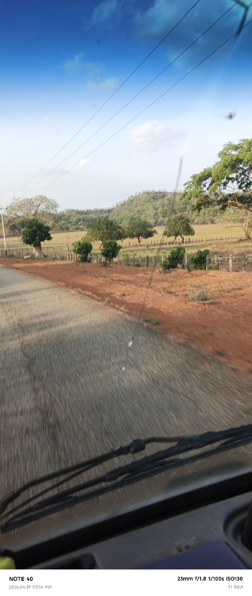 The image, captured from inside a vehicle, possibly in motion, shows a view of a rural landscape near La Cruz, Venezuela. The foreground features the vehicle's dashboard and windshield wipers, slightly blurred, implying movement. Outside, a paved road runs alongside a reddish-brown strip of land before transitioning to a grassy plain. Trees are scattered along a fence line that separates the road from a field that ends towards a green, hilly horizon. The sky above is partly blue with a few wispy clouds, and power lines stretch across the frame. The camera settings at the time of the photograph are noted in the lower right corner.