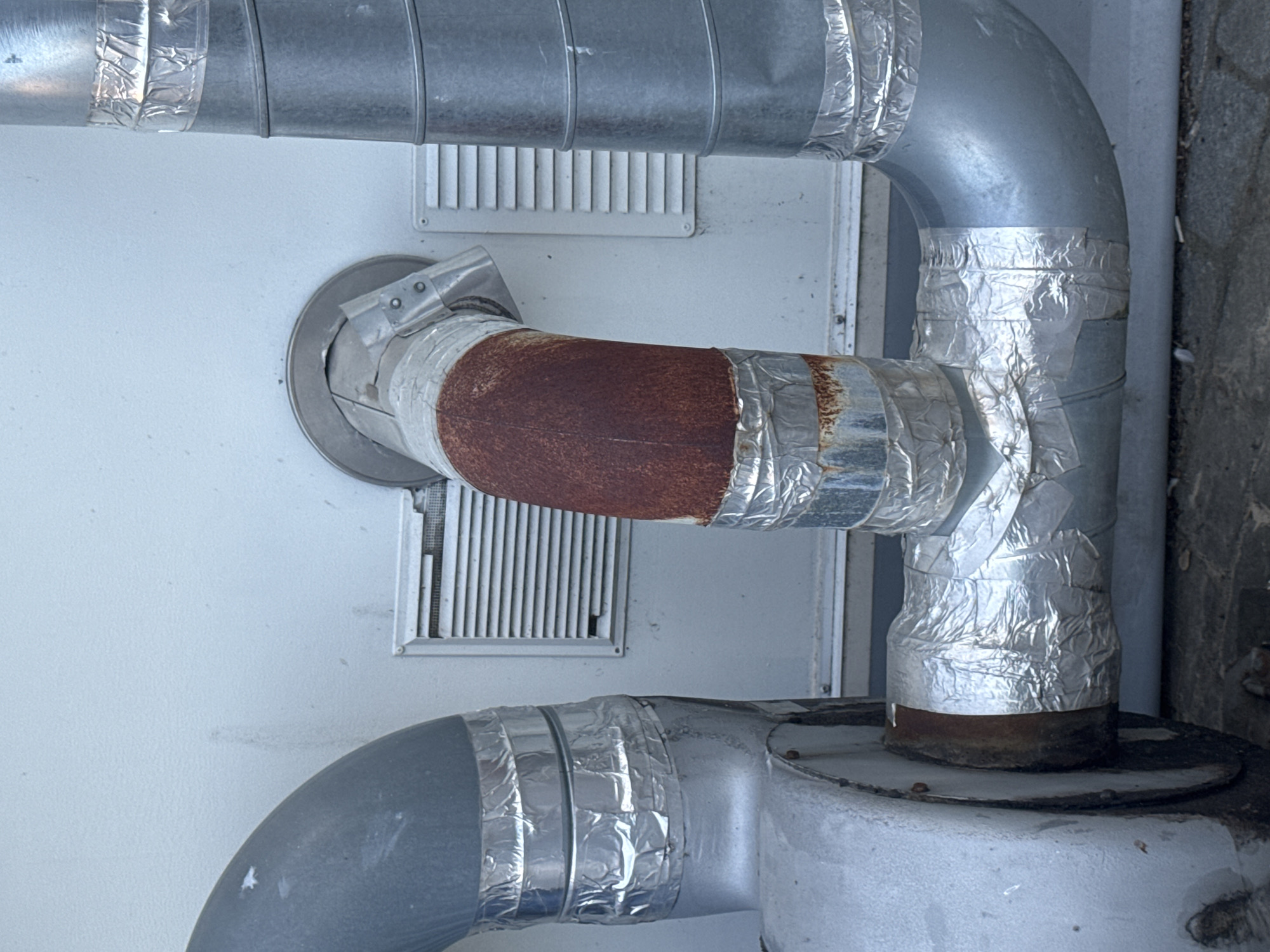 The image shows an exterior view of industrial ventilation pipes. The pipes are made of metal, with some sections showing signs of rust and wear. They are insulated with reflective foil tape in several places. Two rectangular ventilation grilles are visible on a light-colored wall behind the pipes. The scene appears to be located in an urban or industrial setting. The image was taken in Strombeek-Bever, Belgium.