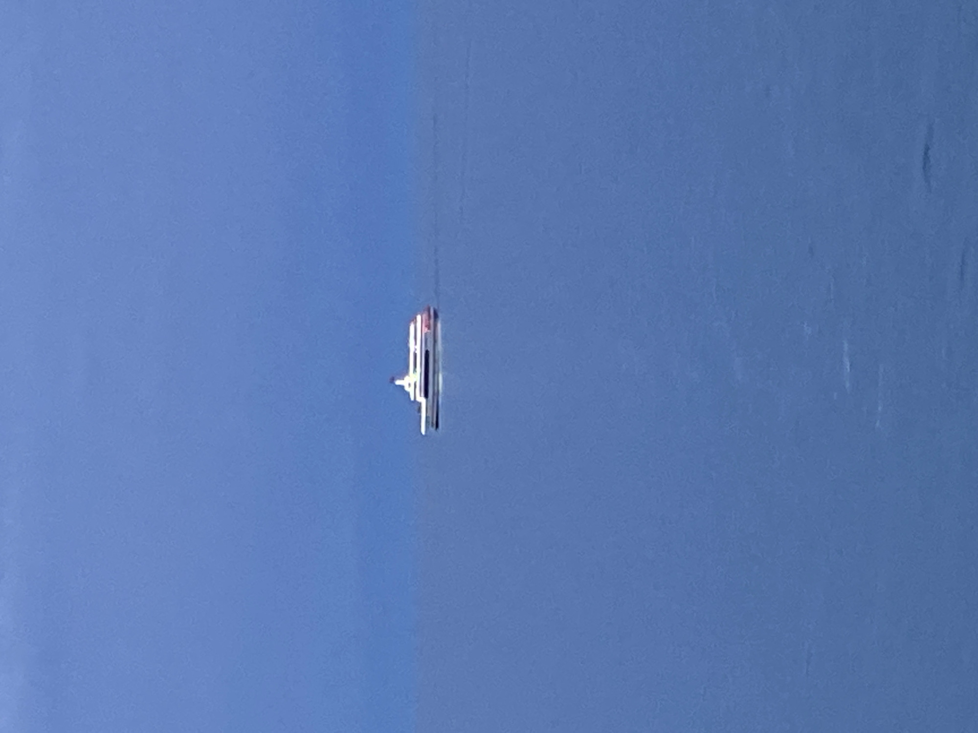A distant view of a white ferry with red accents is depicted on a calm, blue body of water under a hazy sky. The ferry is positioned slightly off-center to the right. The vast expanse of the water and the muted colors of the sky and sea suggest a peaceful, potentially overcast day. No city landmarks or recognizable structures are visible, making it impossible to determine the specific location within Bosteri, Kyrgyzstan. The focus is solely on the solitary vessel navigating the water.