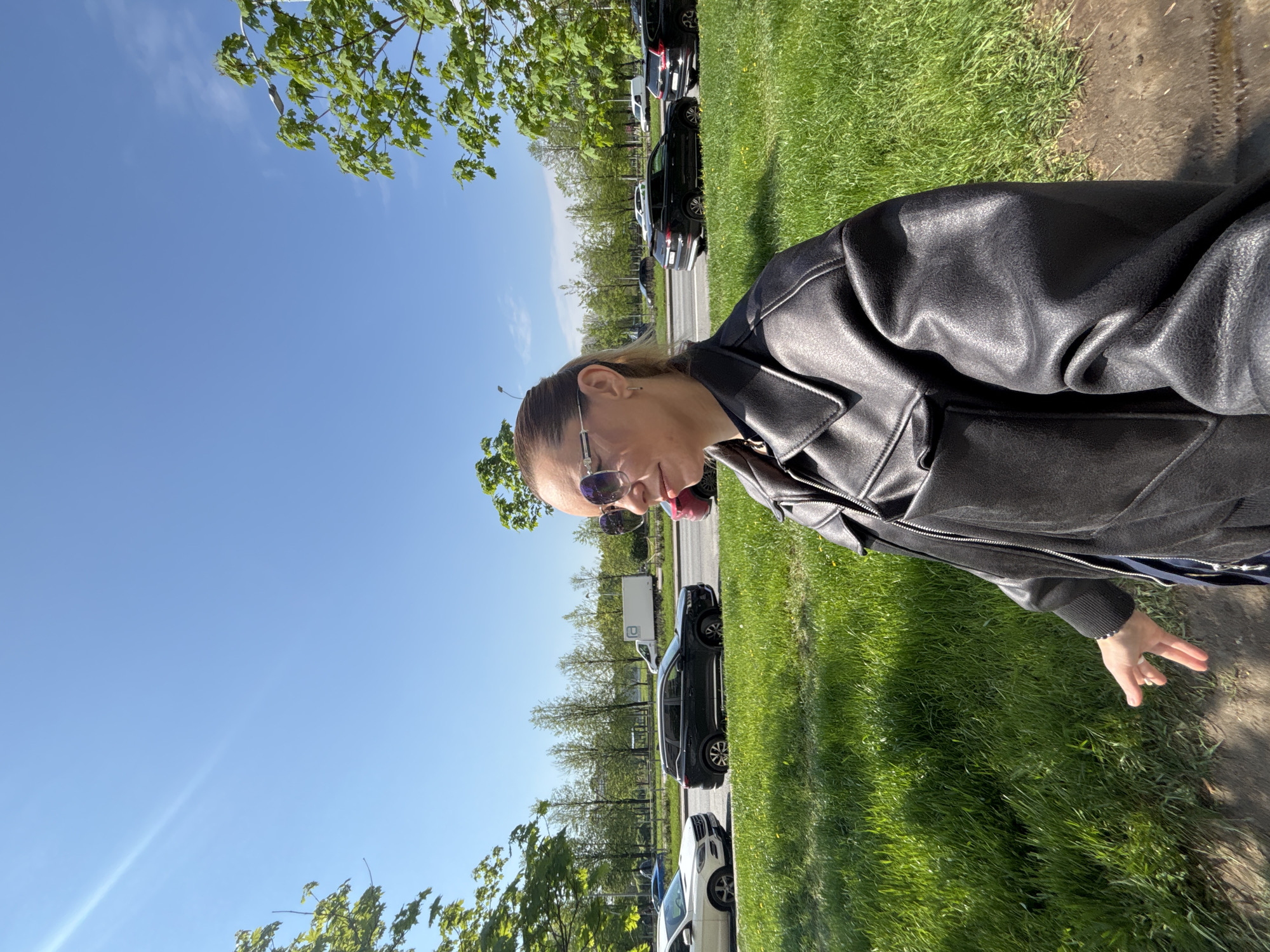 The image shows a woman wearing a black leather jacket and sunglasses standing on a grassy area next to a road. Several cars are parked along the road. The location appears to be in a residential area with trees and a clear blue sky. The city is Vsevolozhsk, Russia.