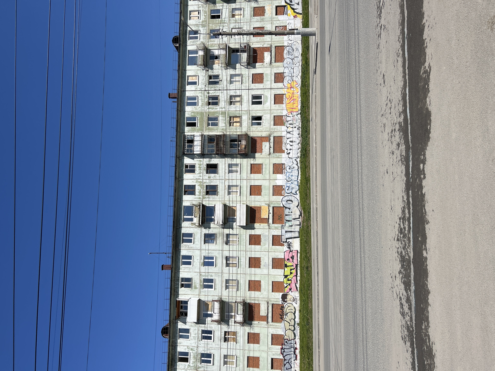 The image depicts a weathered, multi-story residential building in Izhevsk, Russia. The building is pale green and shows signs of disrepair, with boarded-up windows and crumbling facade. Graffiti covers the base of the building along the sidewalk. A paved road runs parallel to the building. Power lines stretch across a clear blue sky above. No people are visible in the frame. The overall scene presents an impression of urban decay.