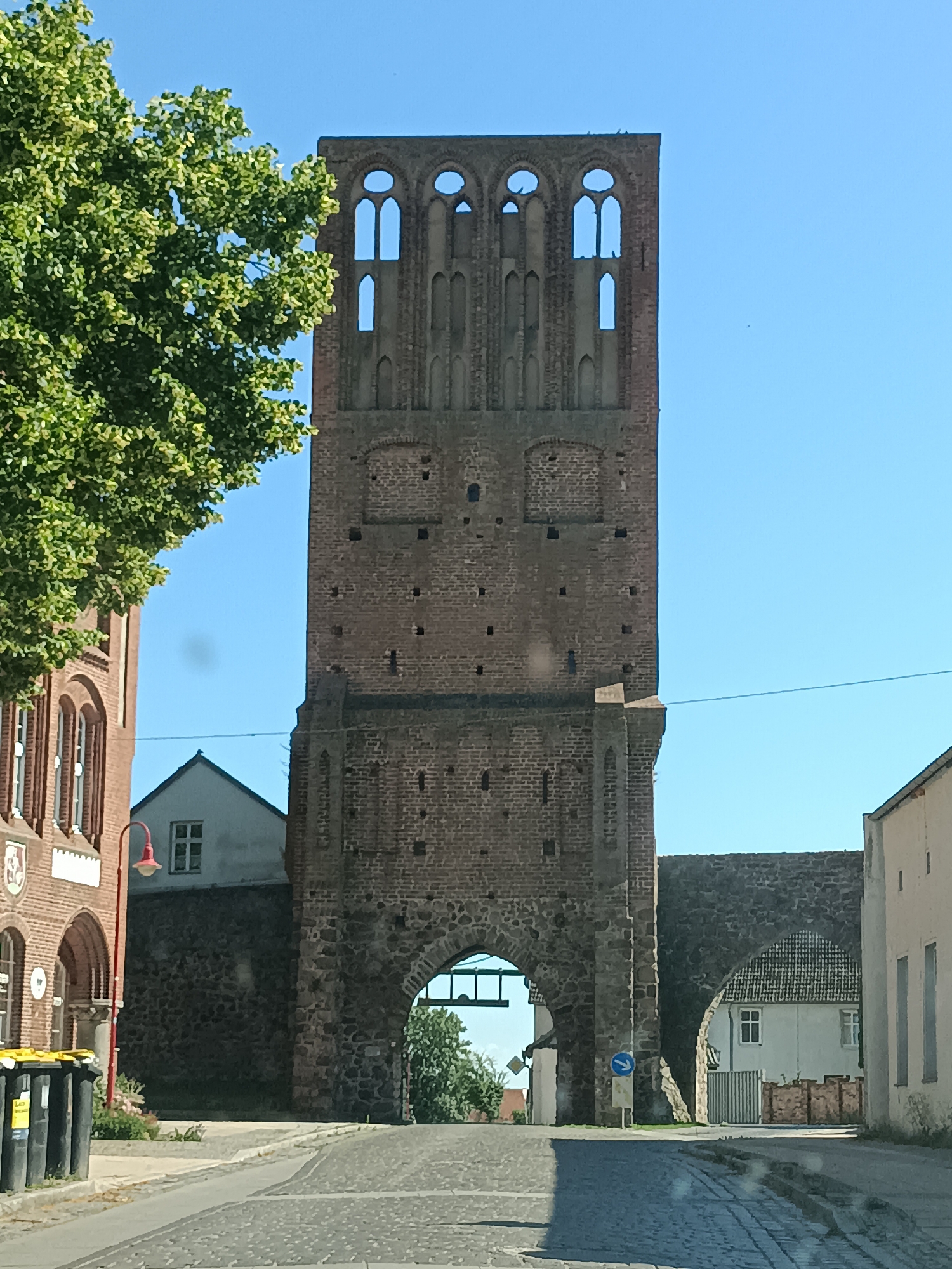 This is a photograph taken from a low vantage point, looking up at the medieval stone gate tower in Gartz (Oder), Germany. The tower is tall and made of dark red brick, with a large arched opening at its base. The upper portion of the tower has multiple arched windows, some of which are open, revealing the clear blue sky beyond. A large green tree with dense foliage is visible to the left of the tower. To the left of the tower, a portion of a red brick building with arched windows is visible, and a red lamppost stands in front of it. In the foreground, a cobblestone street curves towards the gate tower. To the right of the tower, a stone wall with an arched opening leads to a residential area with white houses. The scene is bathed in bright sunlight, suggesting it is daytime. There are no people visible in the image.