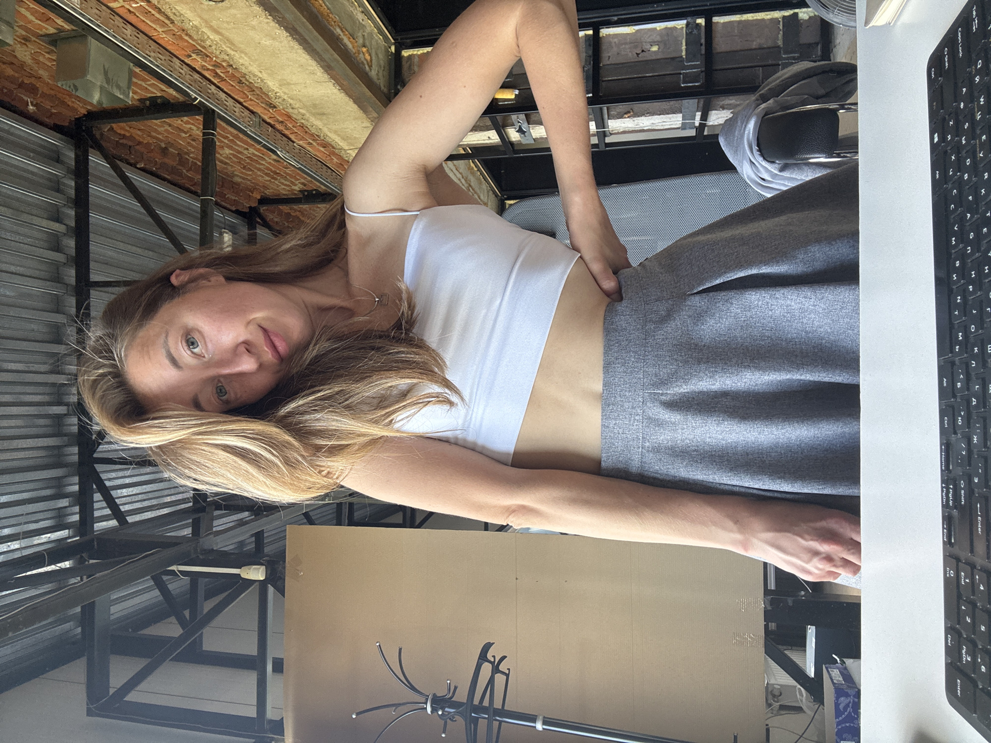 A young woman with blonde hair is leaning towards the camera, her face tilted slightly. She is wearing a white crop top and a grey skirt. Her left arm is bent at the elbow, and her hand rests on her hip. Her right arm is extended, with her hand resting on a desk. A keyboard is visible on the desk to the right. The background appears to be an indoor industrial-style setting with exposed brick and metal structures. There is a large cardboard panel leaning against a wall behind her. The lighting suggests it could be daytime.