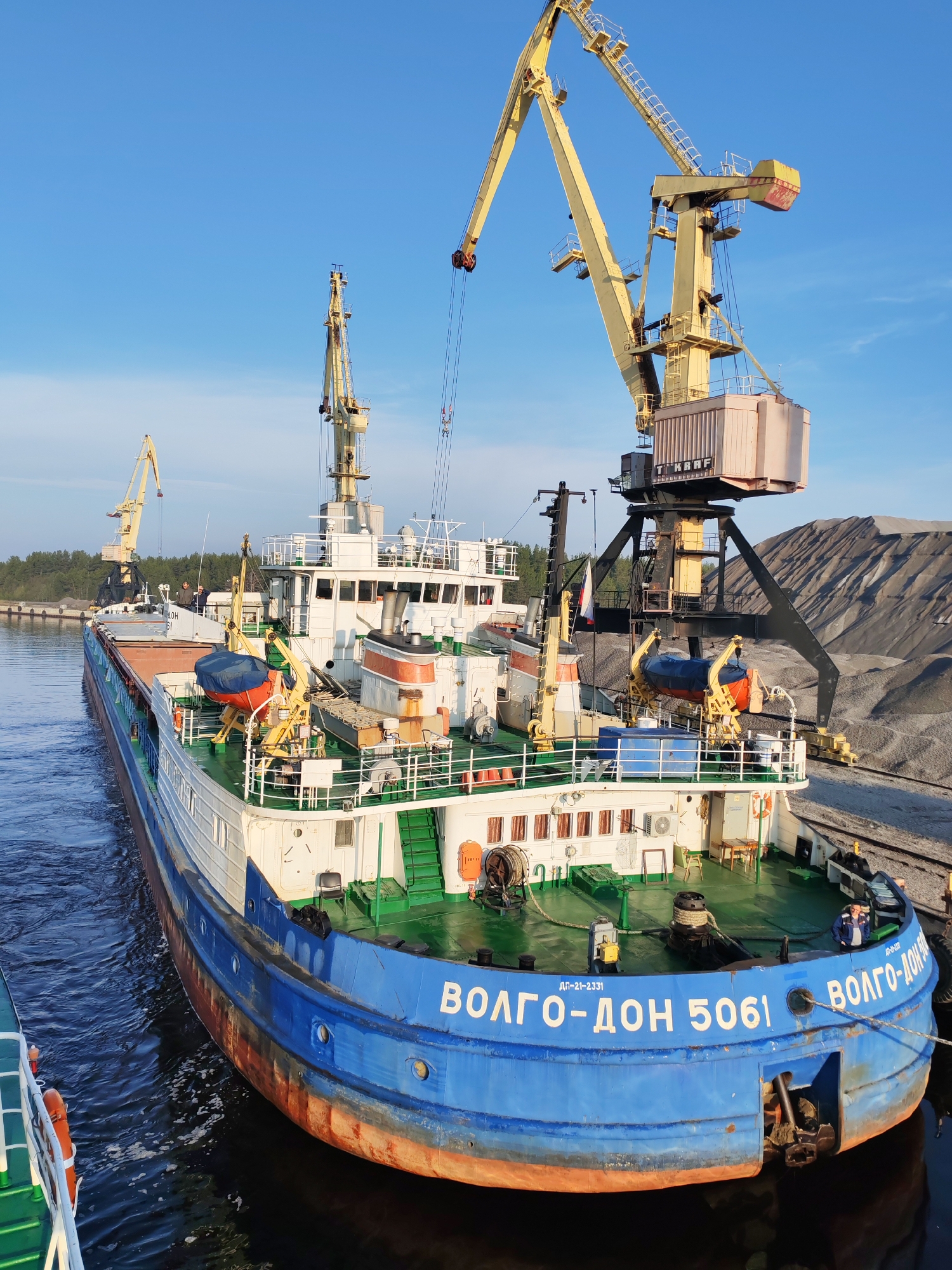 A large cargo ship named "Volgo-Don 5061" is docked in Shal'skii, Russia. The ship is primarily blue and white, with some orange rust along the lower hull. Two large yellow cranes are positioned next to the ship, with one appearing to be in operation. Piles of gray material, likely raw cargo, are on the dock behind the ship. The sky is clear blue. Some trees are in the background, suggesting a coastal or riverside environment. A small portion of another green ship is visible on the lower left.
