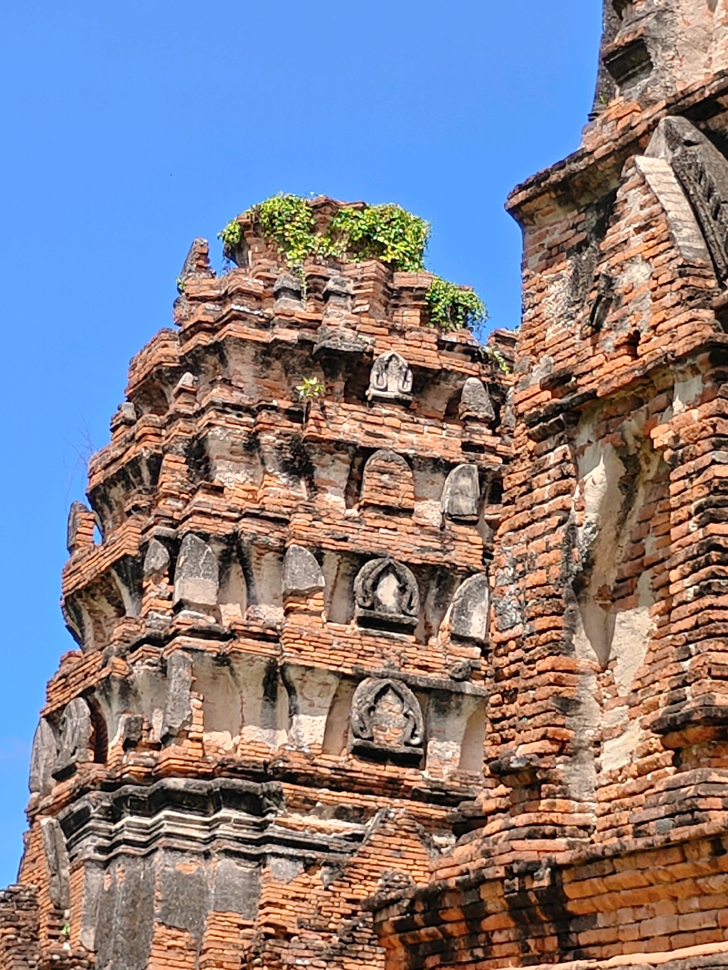 The image shows a close-up view of the weathered brickwork of an ancient temple ruin, with a clear blue sky in the background. The temple features intricate architectural details, including layered brick structures, decorative niches, and some patches of greenery growing on top. The brickwork exhibits signs of age, with some areas appearing darker and more eroded than others. The scene suggests a historical and possibly sacred site. This appears to be Wat Phra Si Sanphet, a prominent temple in Phra Nakhon Si Ayutthaya City Municipality, Thailand, known for its impressive chedis.