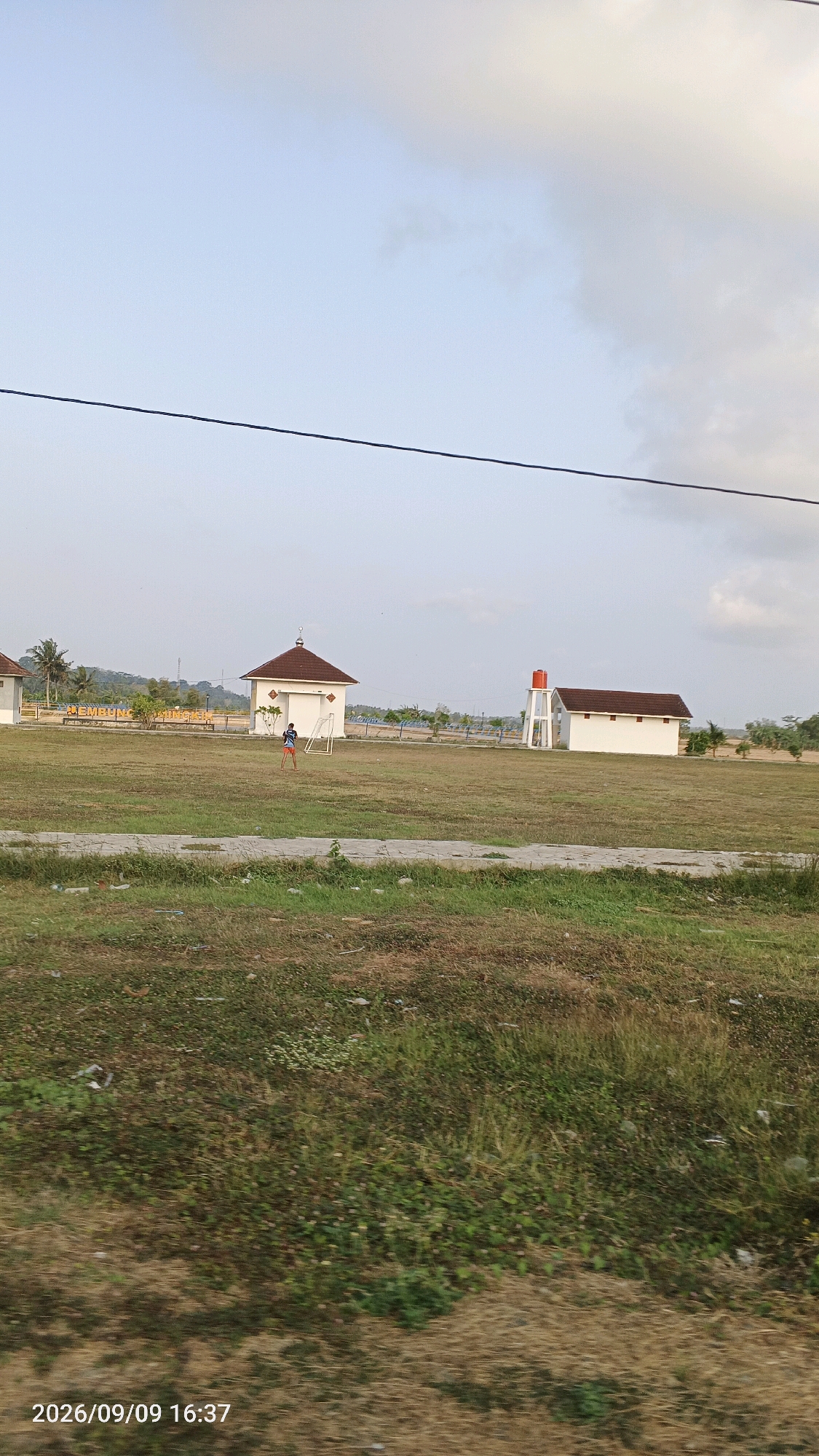 The image depicts an outdoor scene in Sumingkir, Indonesia, featuring a grassy field with several structures. A young person, likely a male wearing a blue shirt and shorts, is visible on the field, positioned near a white structure with a brown, domed roof and an open doorway. A white soccer goal is situated in front of this building. To the right of this building, another white structure with a brown, gabled roof is present, accompanied by a tall, orange water tank. In the background, a sign with yellow lettering is partially visible, reading "NEMBUNG." Palm trees and other vegetation are scattered throughout the landscape, and hills are visible in the distance. The sky is partly cloudy. The timestamp "2026/09/09 16:37" is displayed at the bottom left of the image.