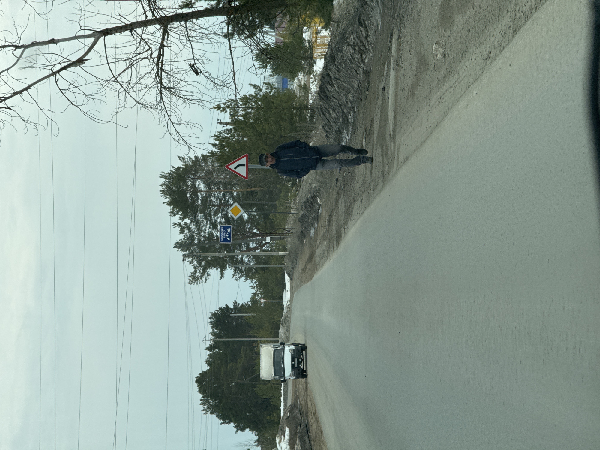 A man in a dark jacket and pants walks along a roadside. A white truck is driving towards the viewer on the road. Several road signs are visible. The location is near Noviy, Russia.