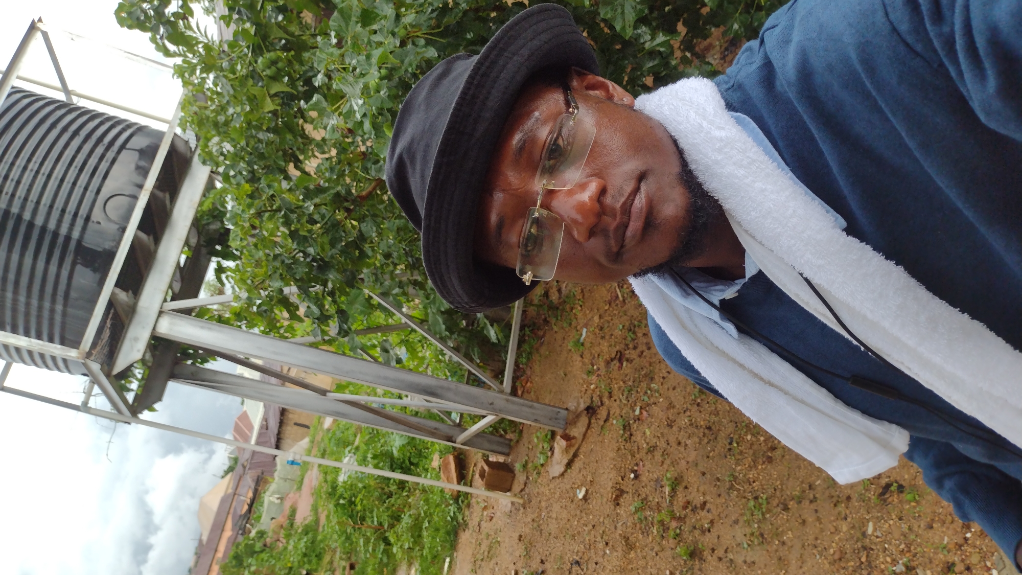 In Jos, Nigeria, a selfie is taken by a man wearing a black bucket hat, glasses, a blue long-sleeved shirt, and a white towel draped around his neck. He has a beard and is looking at the camera with a neutral expression. Behind him, a large black water tank stands on a metal support structure. The background consists of lush green foliage, brown dirt ground, and some buildings in the distance. The sky is overcast with clouds.