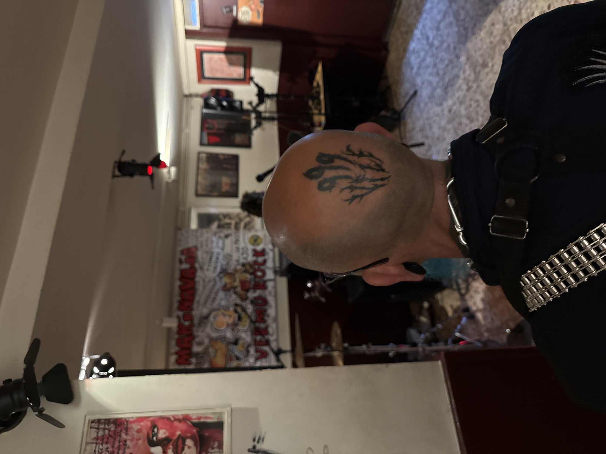 A bald man with a tattoo on his head stands in a room. He is wearing a black shirt and a silver chain necklace. Behind him is a band setting up on stage, with a poster on the wall. The room appears to be a bar or club. Given the poster with "MAK-NAVAJA VERMO ROCK" which is a music venue in Barcelona, Spain.