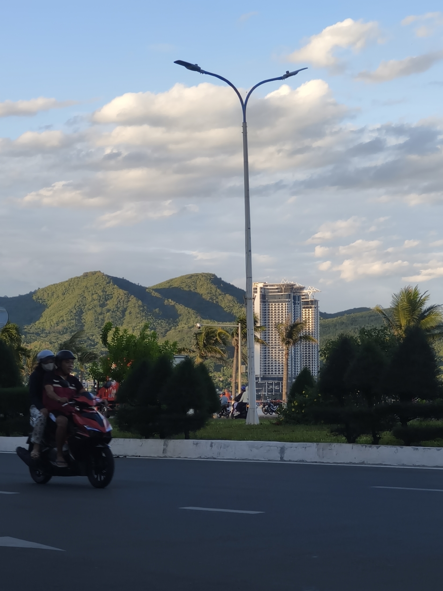 In Nha Trang, Vietnam, two individuals ride a red scooter on a paved road with white lane markings. The passenger in the rear wears a white helmet, while the driver wears a dark helmet and a face mask. Both are dressed casually. The scooter is captured in motion, with a slight blur indicating movement.
Behind the road, a median strip features manicured dark green bushes and some lighter green trees. Beyond the median, a modern high-rise building with a white facade and multiple windows stands prominently. Palm trees are interspersed throughout the landscape, suggesting a tropical or subtropical climate.
In the background, lush green mountains rise, their slopes covered in dense vegetation. The sky above is a clear blue, with scattered white clouds. A tall, slender street lamp with a curved arm extends upwards on the left side of the frame, its silhouette contrasting against the sky. The overall scene depicts a common urban street view in Nha Trang, integrating natural landscapes with modern infrastructure and daily life.