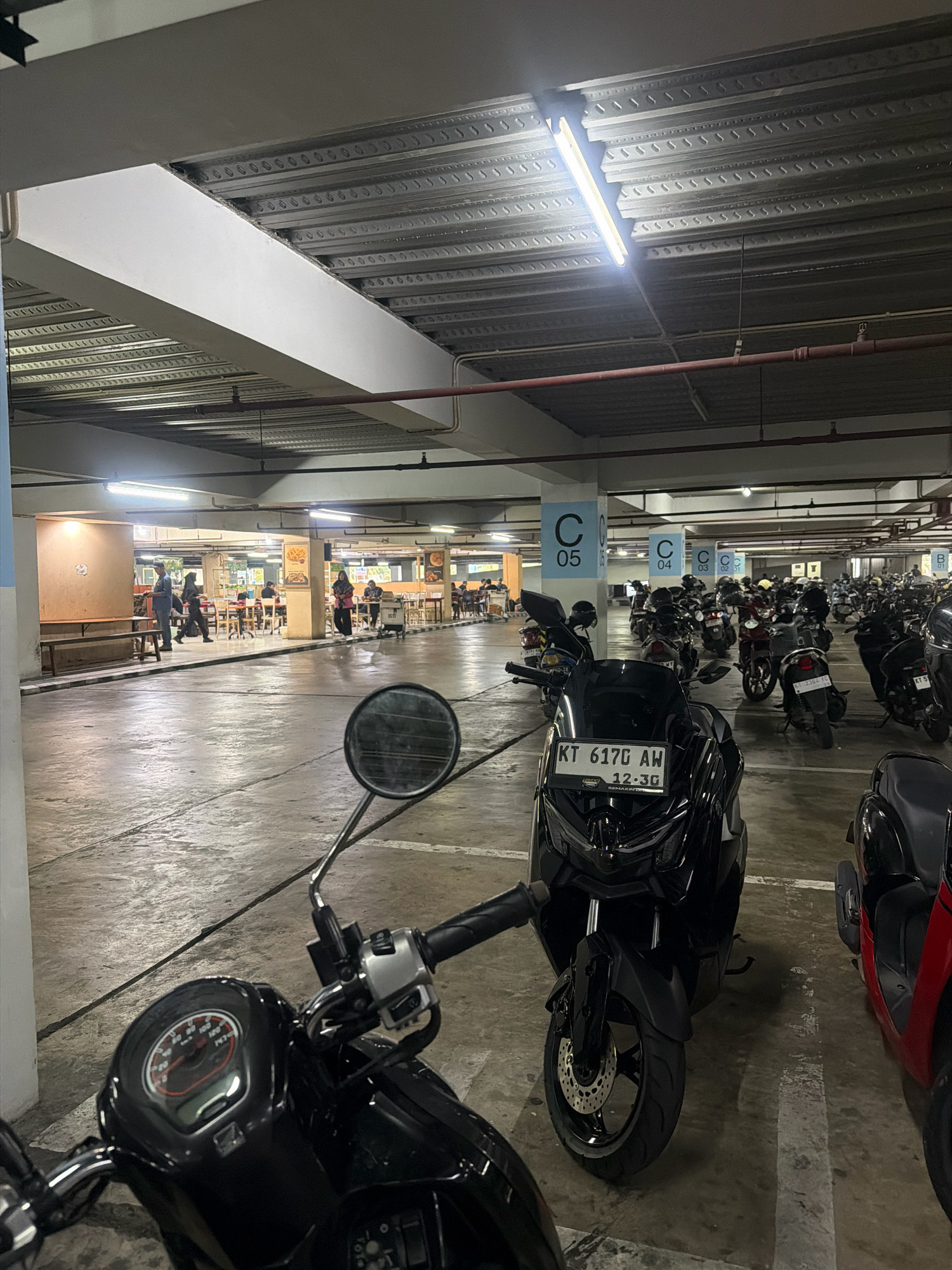 The image depicts an indoor parking area, likely a multi-story car park, in Balikpapan, Indonesia, as suggested by the license plate "KT 617G AW". The ceiling is a ribbed metal structure with fluorescent lighting. The floor is concrete, showing some wear and tear. In the foreground, two motorcycles are parked, with a black scooter on the left and a black sport-style motorcycle with a visible license plate on the right. The license plate of the sport-style motorcycle reads "KT 617G AW 12.3G". In the mid-ground and background, many other motorcycles are parked. Further in the background, there appears to be a food court or dining area with tables and chairs, and several people are visible, some walking and others seated. The overall atmosphere is that of a functional, public parking facility.
