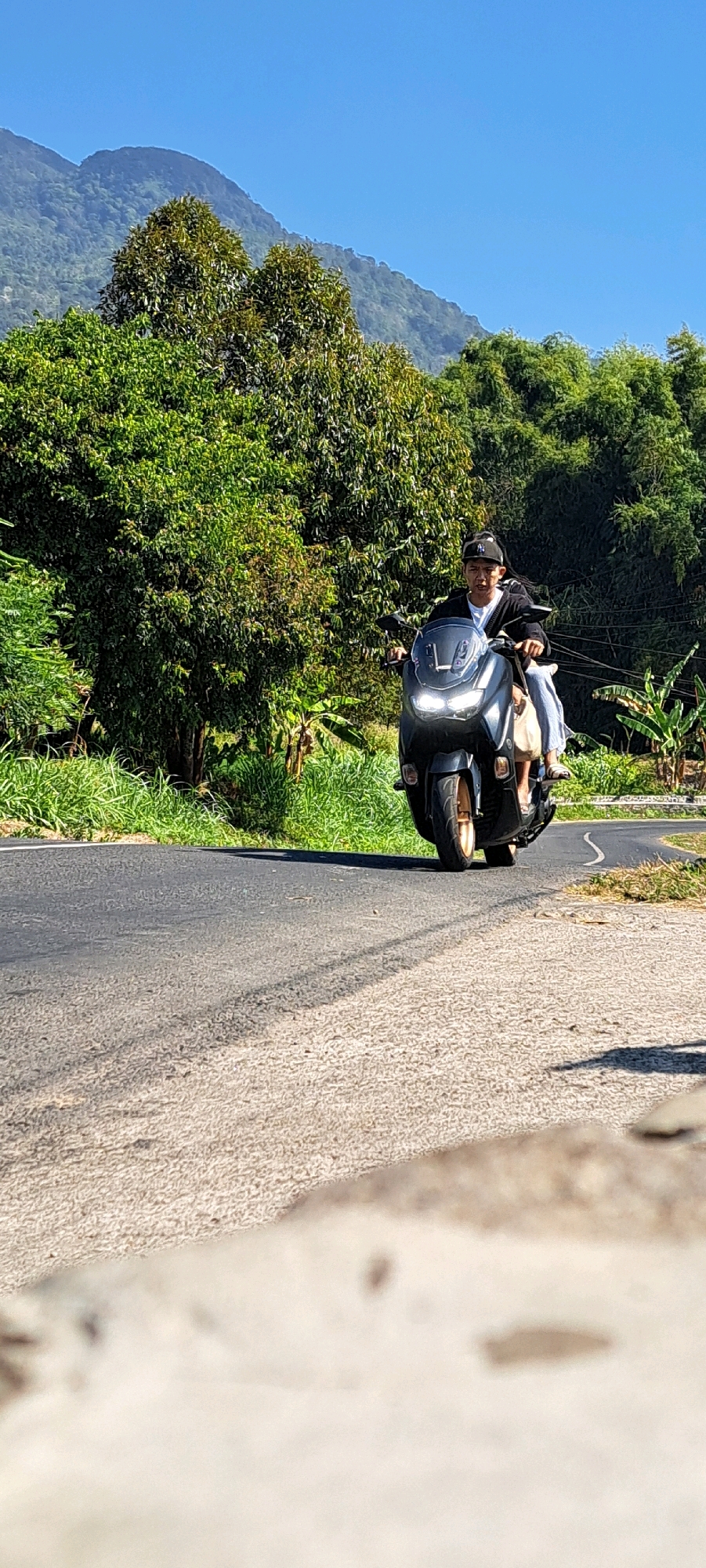 In Sukoreno, Indonesia, a person wearing a black cap and a black jacket over a white t-shirt rides a dark-colored scooter on a paved road. The scooter has its headlights on, and a light-colored bag is visible on the front. The road is bordered by lush green vegetation and trees. In the background, rolling hills covered in foliage are visible under a clear blue sky. The perspective is low, with the foreground of the road being the closest element to the camera.