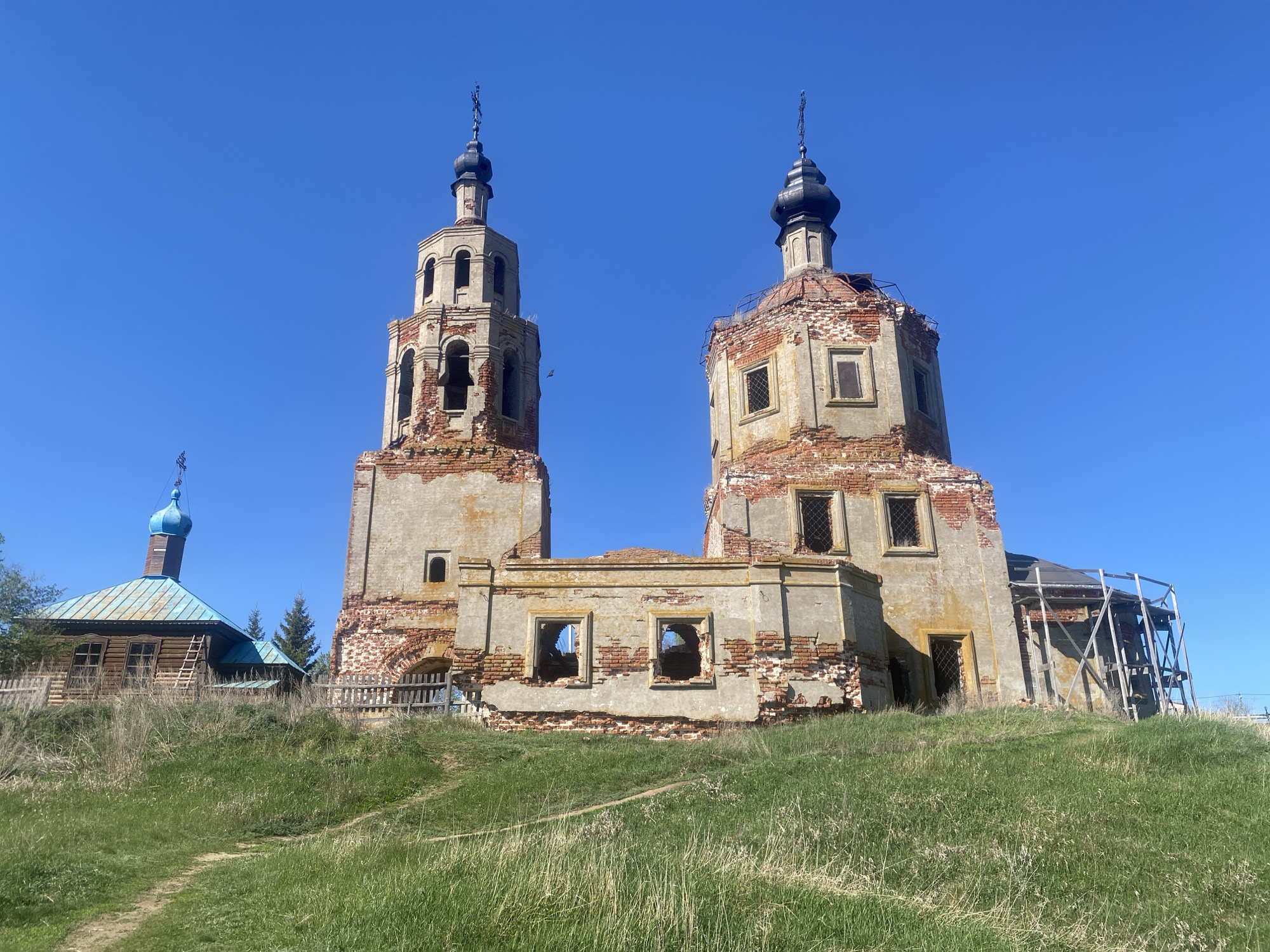 The image shows a partially ruined brick and concrete church in Шуран, Russia. The church has two prominent towers with onion-shaped domes topped with crosses. The structure is heavily weathered and damaged, with exposed brick and missing sections of the outer walls. There are multiple window openings without glass or frames. In the background, to the left of the church, is a smaller wooden building with a blue dome and a wooden fence surrounding the property. The entire scene is set on a grassy hill, with a worn path leading up to the buildings. The sky is clear and blue.