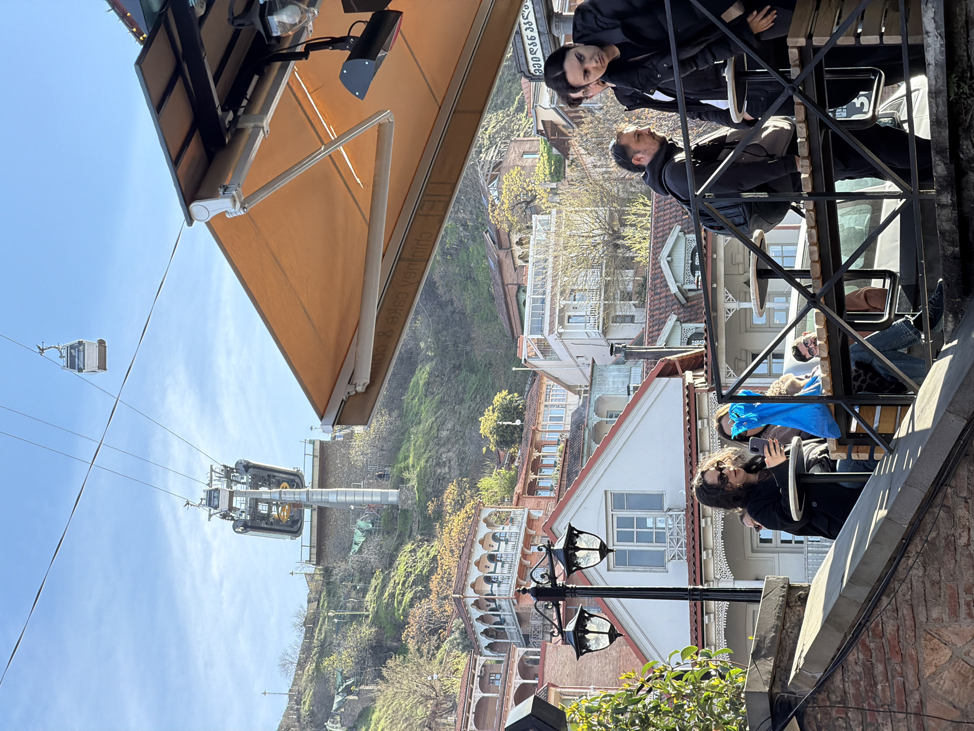 The image captures a scene in Tbilisi, Georgia, featuring a cable car system against a backdrop of traditional architecture and a hillside.
Several people are visible in the foreground, some seated at an outdoor establishment with black metal railings and wooden seating. One individual is holding a smartphone, possibly taking a photo.
In the midground, the landscape is dominated by buildings with distinct red-tiled roofs and white facades, characteristic of Tbilisi's old town. A cable car and its supporting structure are visible in the distance against a clear blue sky.
A tan awning and a street lamp are also apparent in the frame. The environment includes a hillside covered in greenery, adding depth to the urban landscape.
