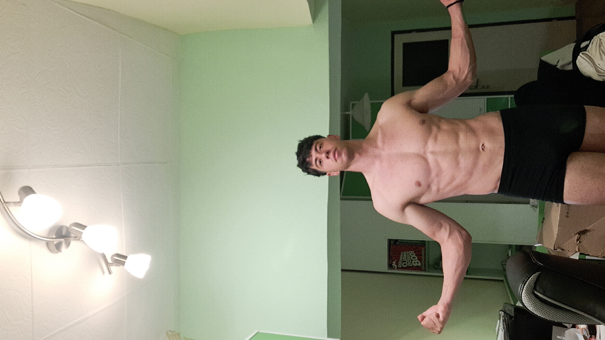 A young, shirtless man with short dark hair is standing in what appears to be a room. He is wearing black briefs and flexing his muscles. The room has light green and white walls. A light fixture with three adjustable spotlights is visible on the white wall. In the background, behind the man, there is a framed picture on the wall, a desk, a chair, and some cardboard boxes. It is not possible to determine if the image was taken in the city of Burgas, Bulgaria.