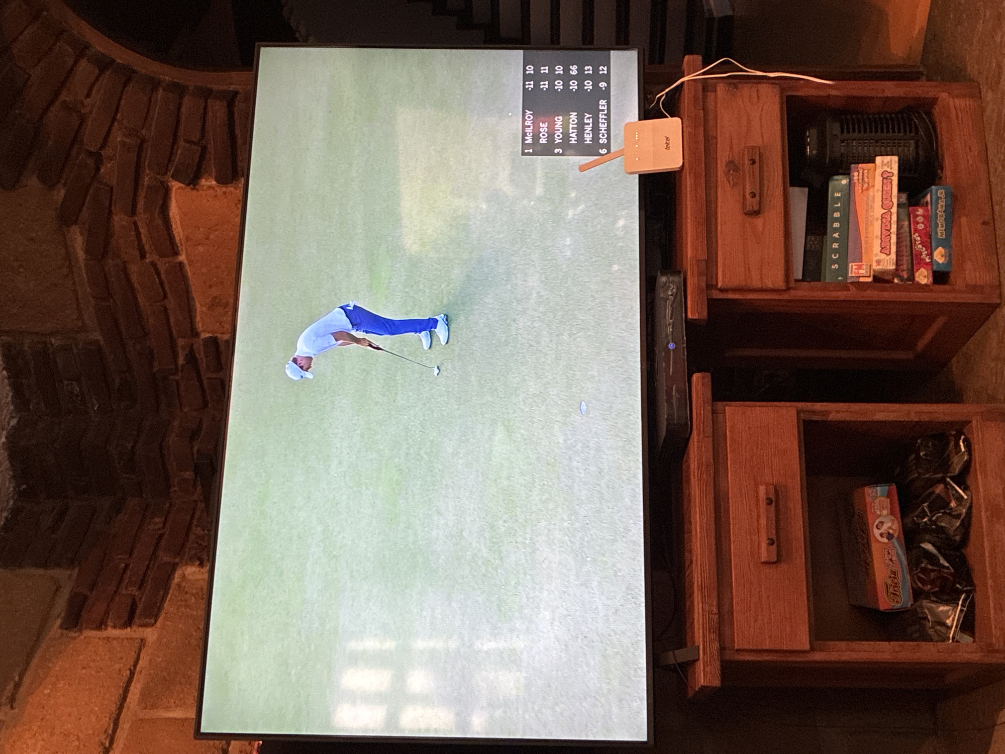 The image shows a television screen displaying a golf tournament leaderboard with McIlroy in first place. A golfer is seen in the middle of a swing on the screen. To the right of the TV are two wooden side tables, one stacked on top of the other. The top table contains board games like Scrabble and Advinia Queen. The bottom table has a Trivial Pursuit game and some other items in dark packaging. The TV is located next to a brick wall, likely a fireplace or structural element. A "Telcel" branded device is connected to the TV. There is no information to suggest the image was taken in the city of Cañada de Madero, Mexico.