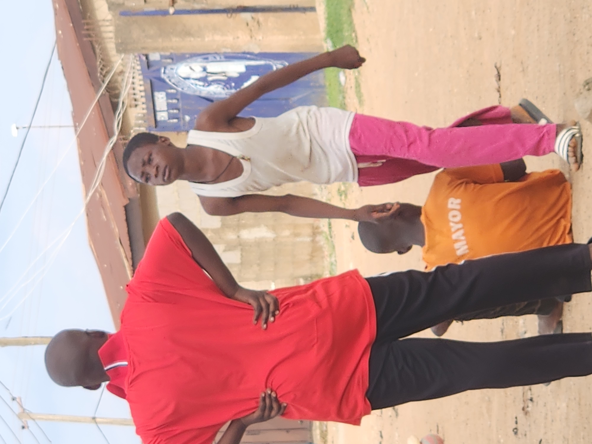 The image depicts an outdoor scene in Sabon Tasha, Nigeria, where three individuals are present in a sandy, dusty environment. On the left, a person wearing a red polo shirt and black pants stands with their hands on their hips, facing away from the camera. In the center and to the right, a young person in a white tank top and bright pink pants is walking, looking to their right. Partially visible in the foreground and to the right of the person in the red shirt is another individual, seen from behind, wearing an orange t-shirt with "MAYOR" written on the back in white lettering. The background features a low-rise building with a corrugated metal roof, a concrete structure, and some power lines overhead. The ground is primarily composed of sand and dirt, with a few scattered pebbles. The overall impression is one of casual, everyday life in an urban or semi-urban setting.
