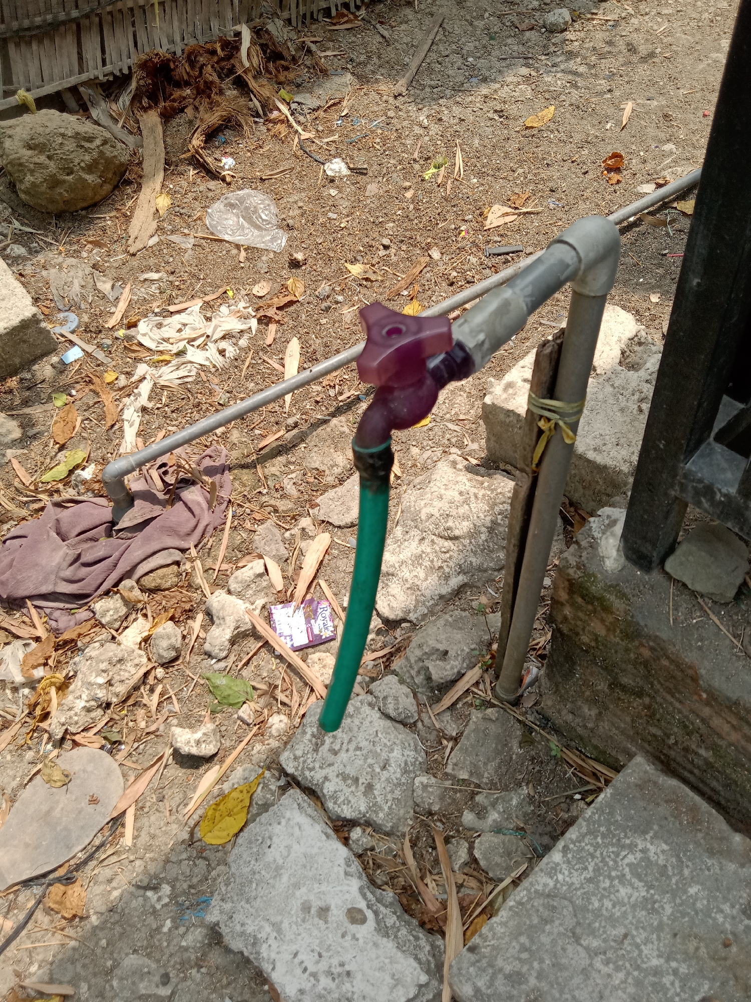 The image shows a water tap with a green hose attached, connected to a pipe system. The tap has a purple handle and is mounted on a grey pipe. The surrounding area is unkempt, with dirt, gravel, scattered leaves, and various pieces of debris including plastic waste and what appears to be a crumpled paper wrapper. There are also some rocks, a piece of brown fabric, and a segment of bamboo fencing in the background. The scene is outdoors, with sunlight creating shadows. The overall impression is of an untidy outdoor area, possibly a yard or alleyway.