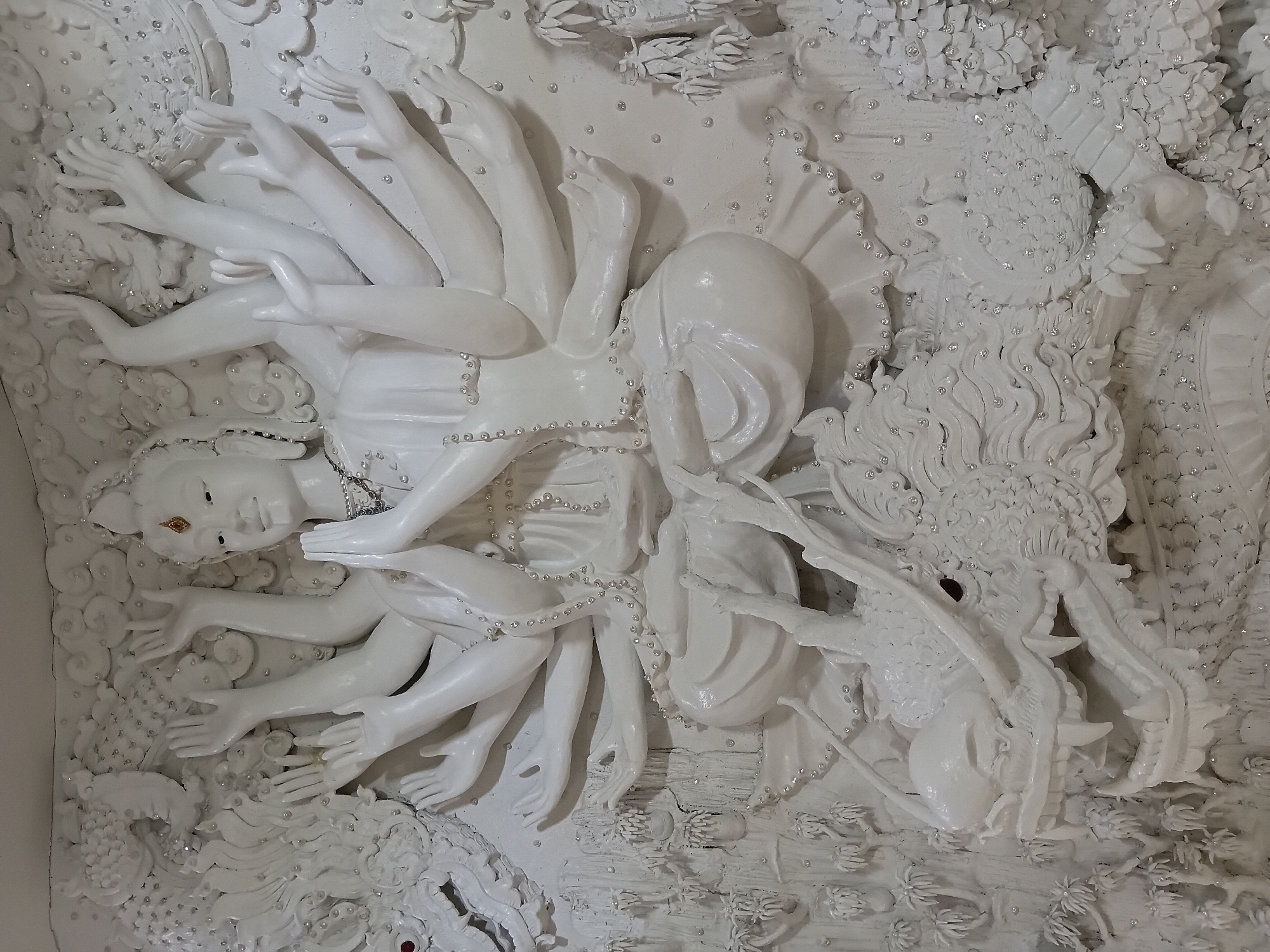 A white, relief sculpture depicts a multi-armed deity sitting in a meditative pose atop a dragon. The deity has numerous arms, some raised and others with hands in prayer or holding objects. The dragon's head is prominent in the foreground, with its mouth open, revealing sharp teeth, and a single red eye. The sculpture is adorned with small, sparkling embellishments that resemble gems. The background is intricately detailed with swirling cloud-like formations and smaller decorative elements, giving the impression of a celestial or mythical scene. The overall style is intricate and ornate. The location of this sculpture is unknown.