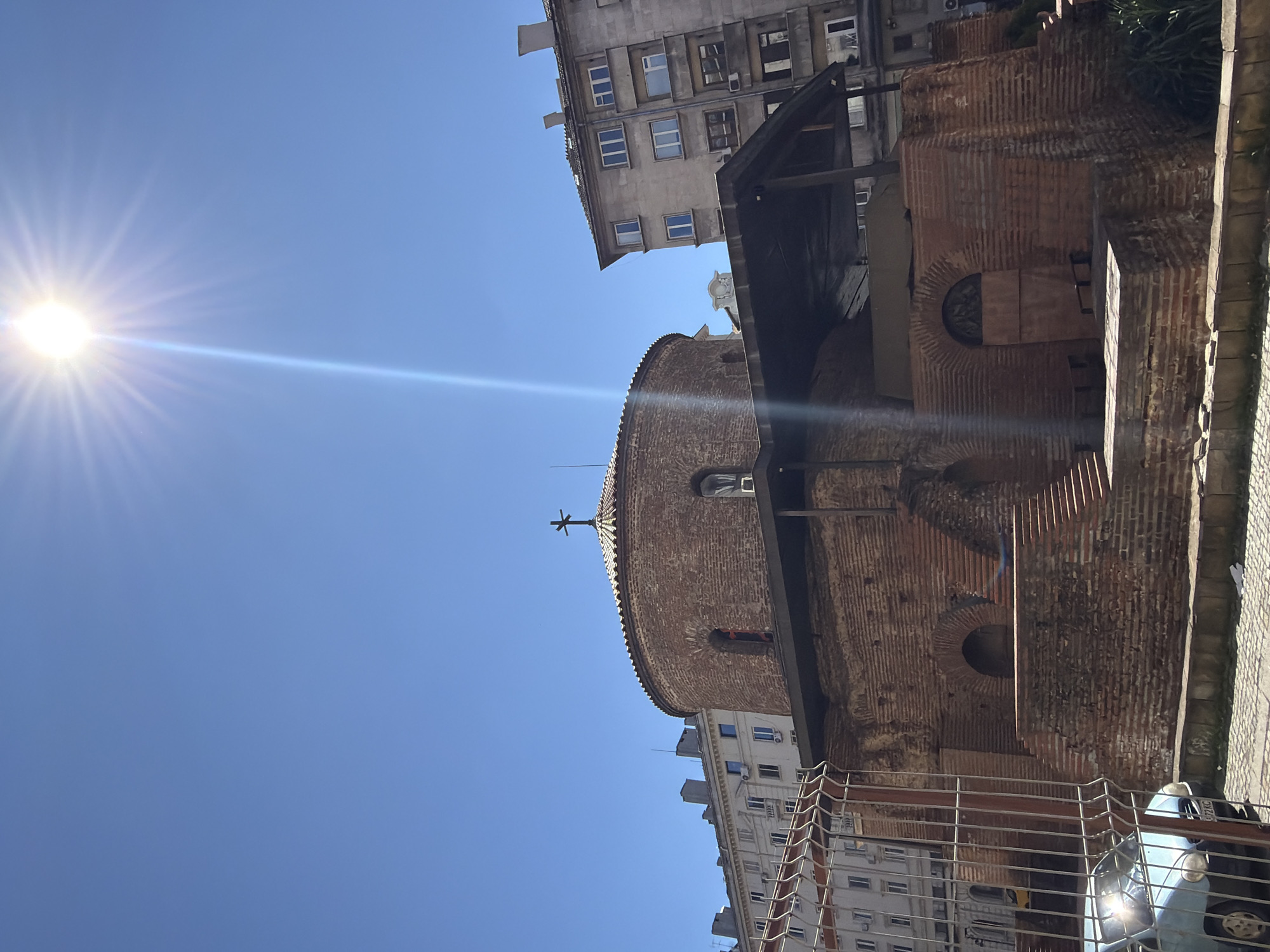 The image depicts the Church of St. George Rotunda in Sofia, Bulgaria, viewed from a low angle. The structure is made of red bricks, with a dome and a cross on top. Adjacent buildings are visible. The sky is clear and blue, with the sun in the upper left corner. A light flare extends across the image. Part of a metal railing and possibly a vehicle are visible in the foreground.