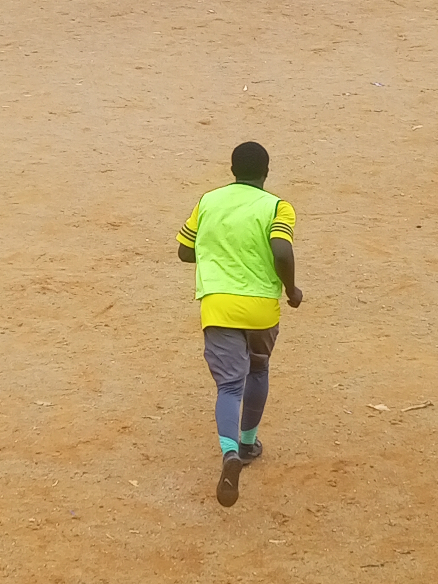 A person is running on a sandy surface. The person is wearing a yellow shirt with black stripes on the sleeves, a bright green vest over the shirt, grey pants with blue and green accents, and dark athletic shoes. The person's back is to the camera, and they are running away from the viewer. The environment appears to be an outdoor area with sparse vegetation and a dirt ground, consistent with a location in Jos, Nigeria. There are no other people or objects visible in the immediate vicinity.