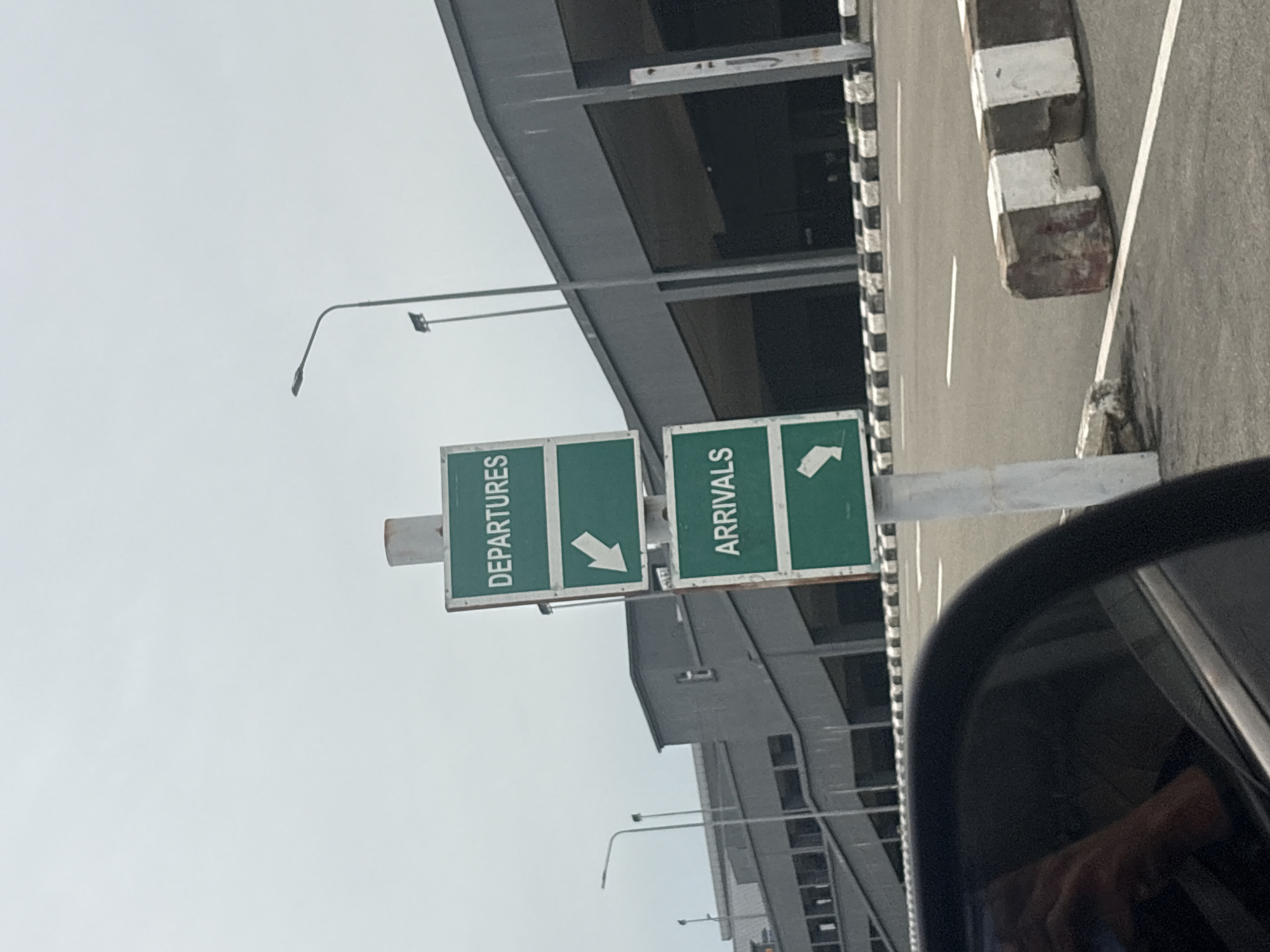 In Gwaska, Nigeria, an airport signage directs traffic. The signs are green with white text and arrows, indicating "DEPARTURES" with an arrow pointing left, and "ARRIVALS" with an arrow pointing down-left. The signs are mounted on a white pole. In the background, a modern airport terminal building with large, dark openings is visible. The sky is overcast. A street runs in front of the terminal, marked with white lines and a black and white checkered barrier. Part of a car's black interior, including a reflection of a hand, is visible in the foreground.