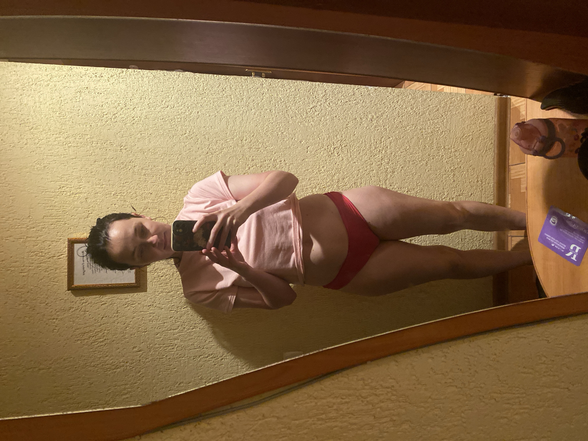 A woman stands in front of a textured yellow wall, taking a selfie in a mirror. She is wearing a pink crop top and red underwear. Her hair is pulled back in a bun. A framed picture hangs on the wall behind her. The mirror shows a reflection of the room, including a wooden floor, a water bottle, and some other objects on a dresser. The overall lighting is dim.