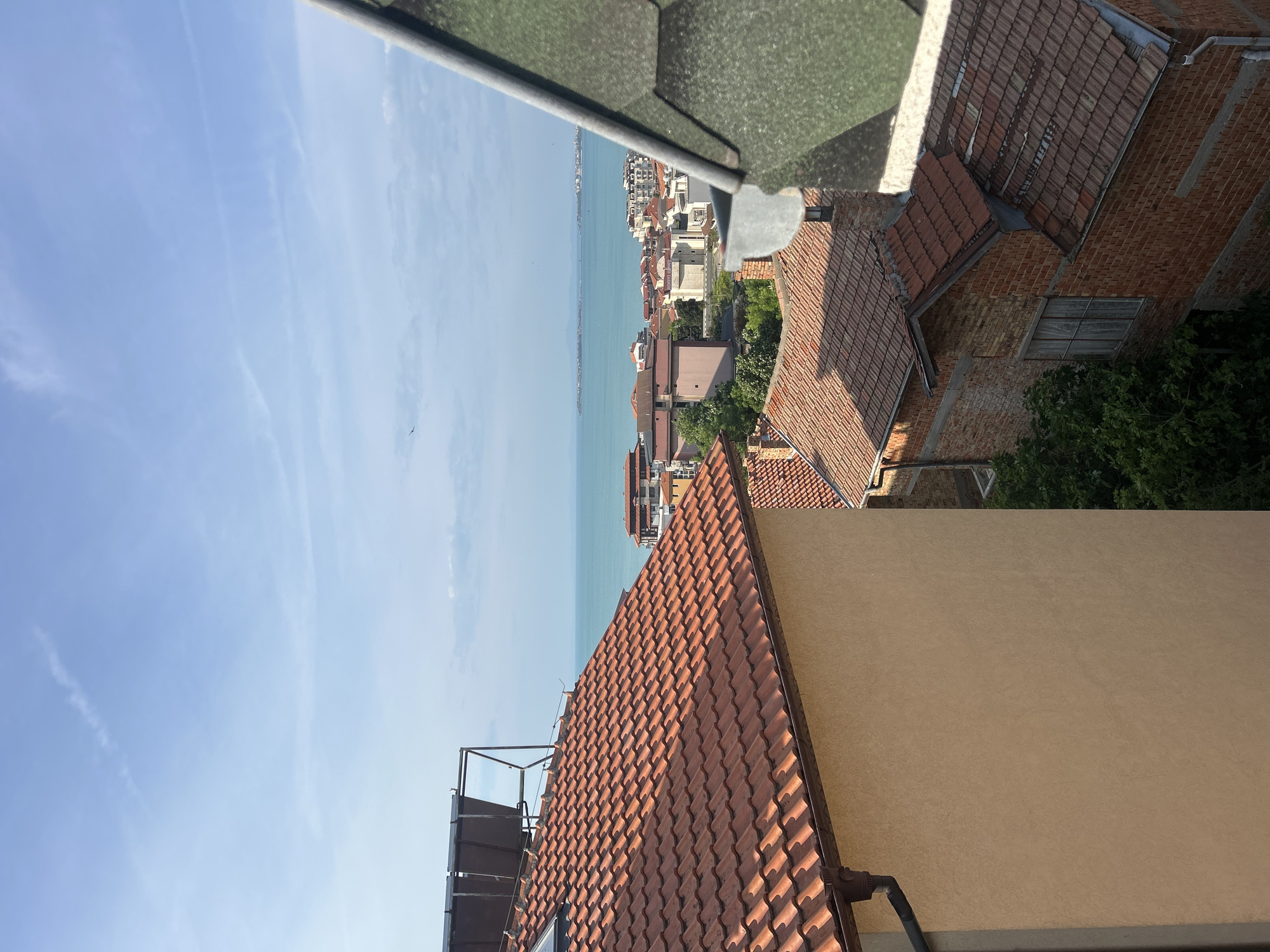 The image shows a view from an elevated position, likely a rooftop in Sveti Vlas, Bulgaria, overlooking a coastal area. The turquoise sea stretches towards the horizon under a light blue sky with faint clouds. The urban landscape of Sveti Vlas includes residential buildings with red-tiled roofs, interspersed with greenery. A building with a red brick facade is also visible. Roofs of neighboring buildings, including one with green shingles, frame the perspective. A section of a building with a tan facade and a satellite dish is visible in the foreground.