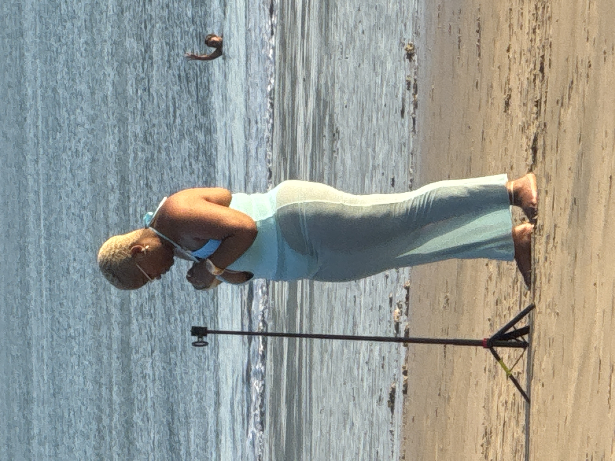 A woman with blonde, cropped hair and sunglasses stands on a sandy beach in Kuta, Indonesia, facing a tripod with a camera. She is wearing a light blue halter-top bikini with a sheer, matching long skirt. Her hands are clasped in front of her chest. In the background, the ocean is visible with gentle waves rolling towards the shore. A person is partially submerged in the water to the right of the frame, with their arm raised in a wave. The lighting suggests it is daytime, likely late afternoon due to the warm tones. The scene captures a moment of apparent relaxation or a planned photo opportunity by the sea.