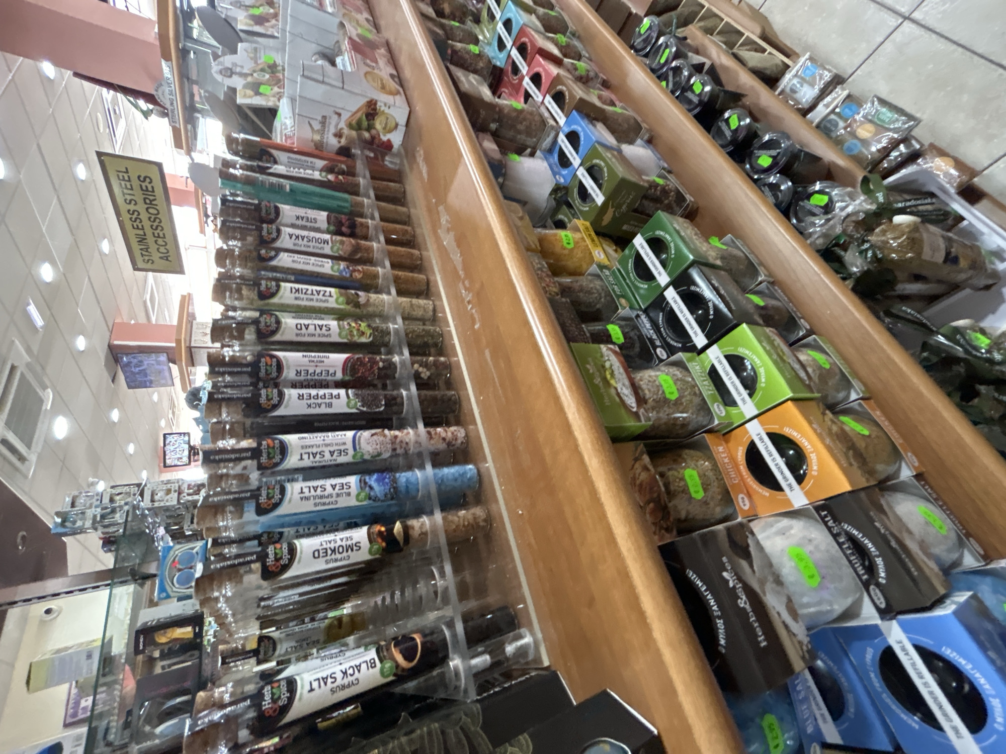 This is a medium shot of a retail store shelf displaying a variety of spices and seasonings. The shelves are made of wood and are filled with glass jars and cardboard boxes containing different types of salts and spice mixes. The products are labeled in English and Greek, with some labels indicating they are from Cyprus. The store appears to be well-lit, with fluorescent lighting visible on the ceiling. The overall impression is of a well-stocked specialty food store. Given the product labels and the general aesthetic, this store is likely located in Ayia Napa, Cyprus.