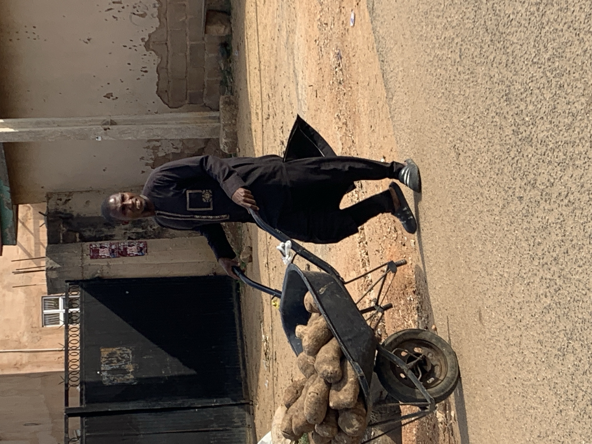 A man in a black outfit is pushing a wheelbarrow filled with yams on a paved road. The location appears to be near a building with a gated entrance. The image was taken in the city of New Stadium, Nigeria.